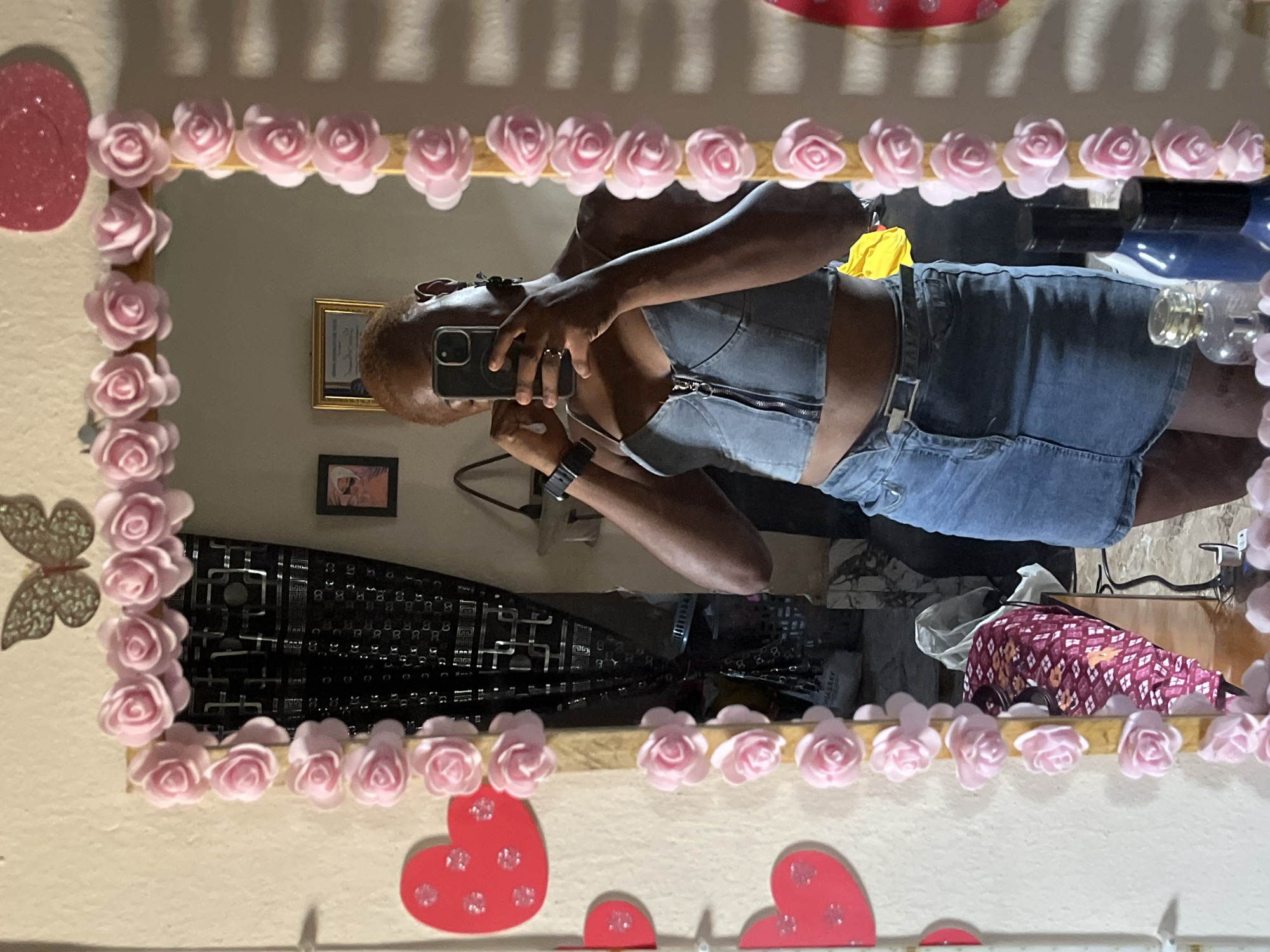 This image is a mirror selfie taken indoors, rotated 90 degrees counter-clockwise. The scene captures an individual, likely an adult with a dark complexion and short, reddish-brown hair, taking a photo of themselves in a mirror. They are wearing a denim crop top featuring a zipper down the front and denim shorts or a skirt. A black watch is visible on their left wrist, and they are holding a silver or grey smartphone, possibly an iPhone, to capture the reflection.
The mirror's frame is distinctly decorated with a continuous border of small, light pink faux roses. On the wall surrounding the mirror, additional decorations include glittery red heart cutouts of various sizes and a glittery butterfly shape. The reflected background reveals a room with light-colored walls, common in residential settings in Jalingo, Nigeria. Visible elements in the reflection include two framed pictures: a larger one with a gold frame and a smaller one with a dark frame, hung on the wall. Other reflected objects suggest a lived-in space, such as a dark object with a grid pattern, a blue bottle, and various other items. The lighting appears to be bright indoor illumination. No discernible text is visible on any of the reflected items.