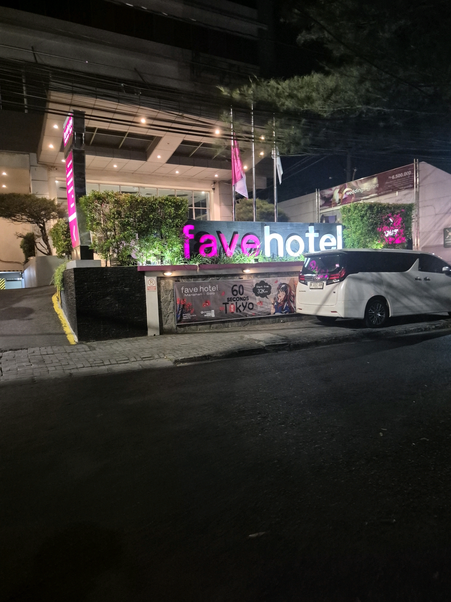 The image depicts the exterior of Favehotel in Surakarta, Indonesia, at night. A large, illuminated sign prominently displays "fave hotel" in neon pink and white letters. To the left of the hotel entrance, a tall, dark sign with pink lettering is visible. In front of the hotel, a white minivan is parked. A banner advertising "60 SECONDS TOKYO" with an anime-style illustration and a price starting from "32k++" is displayed on the lower part of the hotel's facade. Another banner, further to the right, advertises "weddings@fave" with a price of "Rp. 6.500.000,-". The hotel building has a modern facade with a covered entrance area. Green shrubbery and trees are planted around the entrance. The ground in front of the hotel is paved with bricks. The scene is illuminated by artificial lighting from the hotel and its signage.