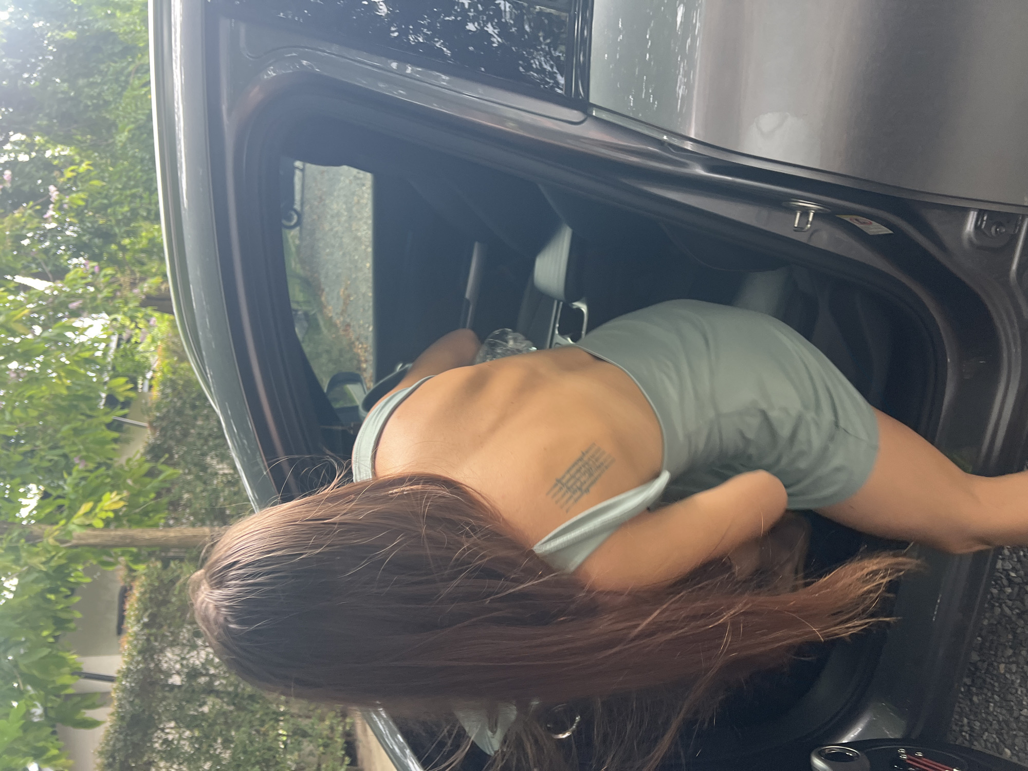 An overhead, slightly tilted image shows a person bending over into the open rear door of a dark-colored car. The individual is seen from behind, their back facing the camera, with long, dark brown hair falling forward as they lean into the vehicle. They are wearing a light green or teal-colored sleeveless top or dress that reveals their upper back. A tattoo with several lines of text or symbols is visible on their upper back, near the right shoulder blade. The script of the tattoo is not clearly discernible.
The scene is set outdoors during daylight hours, indicated by the bright ambient light and the lush green trees and foliage visible in the background, suggesting a natural environment. The car appears to be parked on a paved surface, possibly a driveway or roadside, in บ้านบางรักษ์, Thailand. Inside the car's door frame, a small metallic hook or fastener and a tiny white label with illegible text or symbols can be seen. The person seems to be reaching for an object or accessing the car's interior.