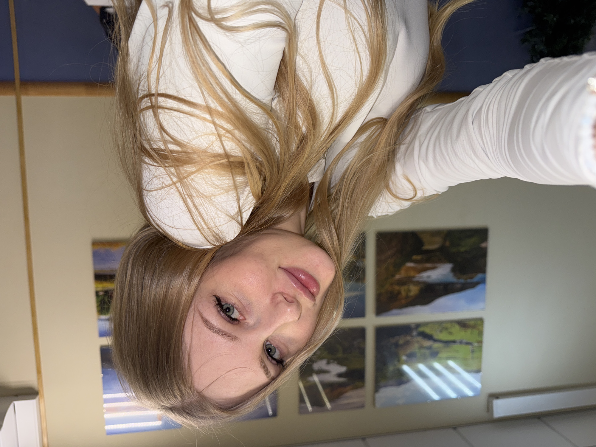 The image is a selfie of a fair-skinned female with long, blonde hair, wearing a white garment with long sleeves. She has visible eye makeup. Behind her, a wall is decorated with four square landscape pictures and a rectangular, white object resembling a vent. The location appears to be indoors. The photograph does not depict the city of Perm, Russia, nor can the location be determined based on the image.
