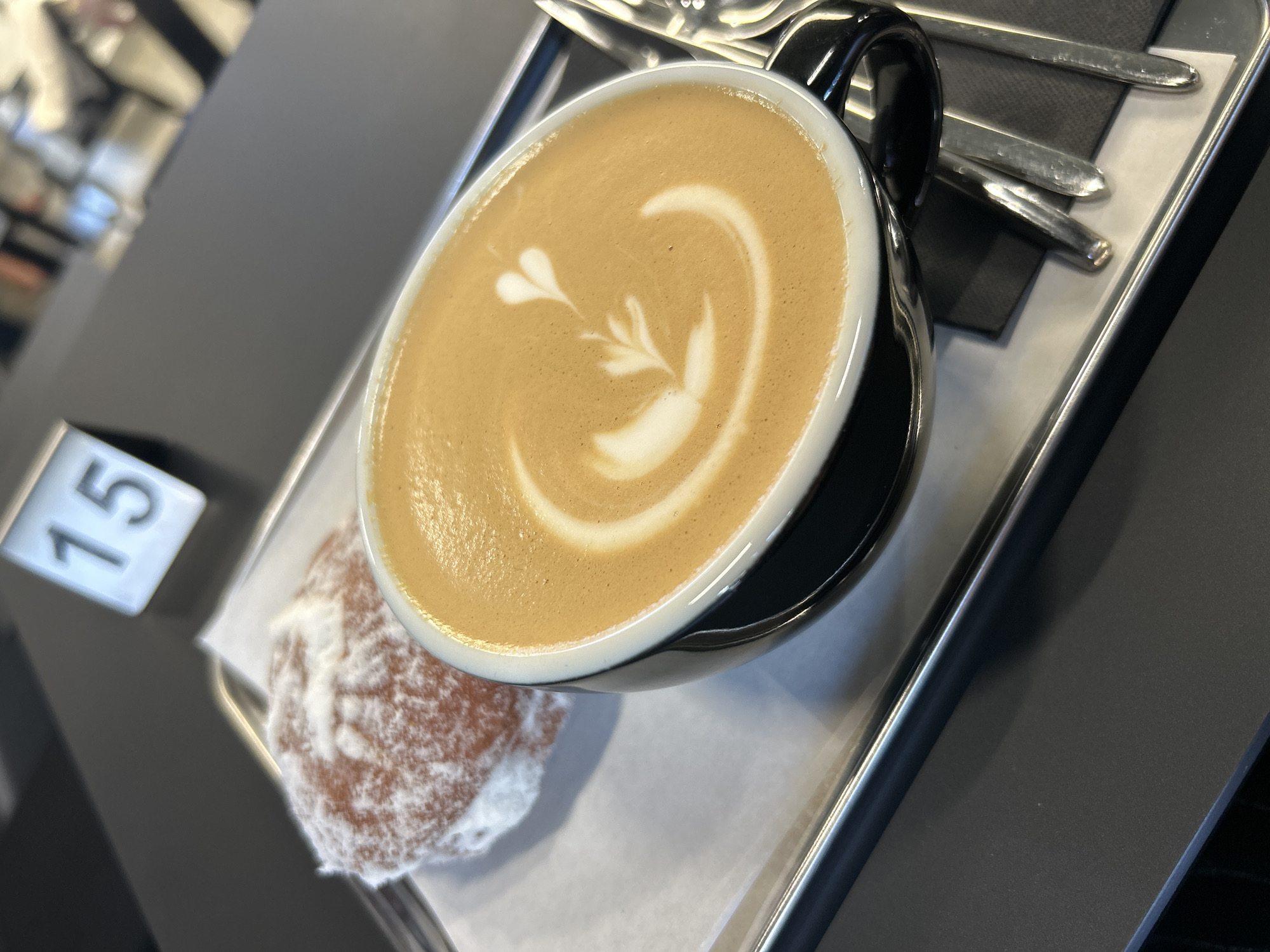 The image presents a close-up of a cappuccino with latte art, accompanied by a powdered doughnut, served on a silver tray. The coffee, in a black mug, features a design in the foam. Utensils are positioned beside the mug on a black napkin. A table number, "15," is visible in the background. The location cannot be determined.