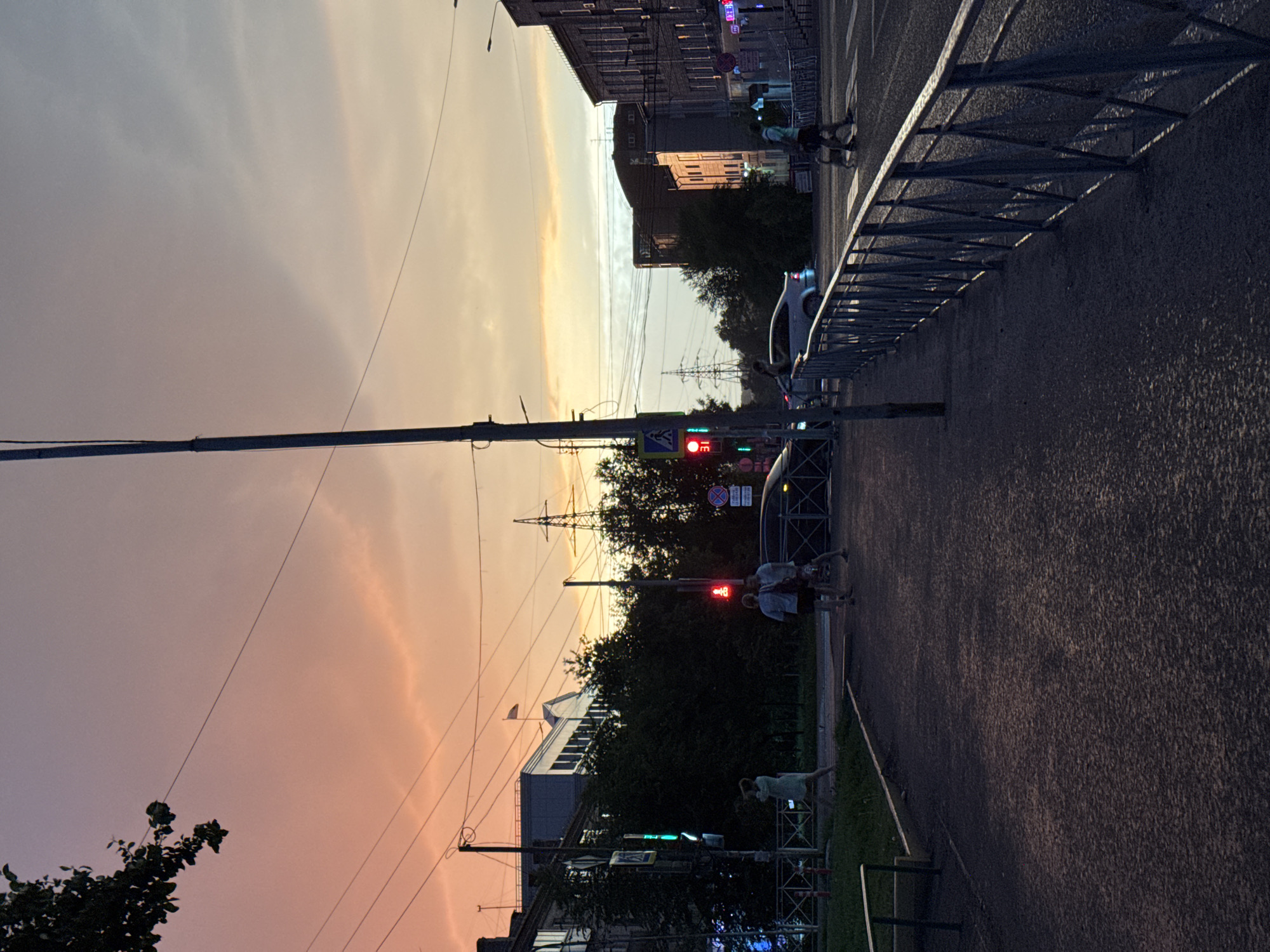 This is a medium-shot, eye-level, vertical photograph taken during sunset in an urban environment, likely in Novosibirsk, Russia, given the visual cues of the architecture and traffic signals. The sky is a prominent feature, displaying a gradient of colors from soft orange and pink near the horizon to a pale blue at the top, with some scattered clouds.
In the foreground, a dark asphalt pedestrian walkway runs along the right side of the frame, bordered by a grey metal railing. The railing casts a shadow on the walkway. On the left side of the walkway, there is a patch of grass and a curb.
In the midground, there is a street with a crosswalk and several people. On the left side of the street, a woman in a light-colored dress is walking. Further down the street, three people are walking near a traffic light that is red. Cars are also present on the street, including a silver sedan and a darker vehicle. A pedestrian crossing sign is visible on the left.
The background features buildings of varying heights and architectural styles, some appearing to be residential or commercial. Trees are scattered throughout the scene, particularly behind the buildings on the left. Electrical poles and power lines are visible crisscrossing the sky, along with a prominent transmission tower in the distance. The overall lighting suggests dusk, with artificial lights from buildings beginning to illuminate.