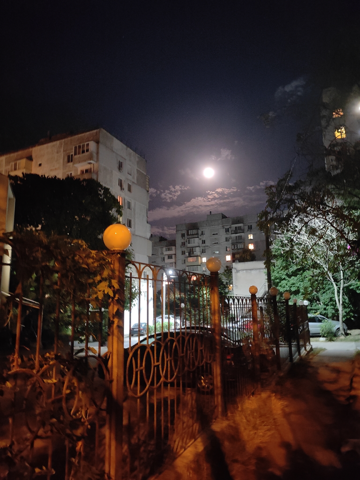 The image captures a nighttime scene in the city of Chornomorsk, Ukraine, with a full moon prominently featured in the dark sky, partially obscured by scattered clouds. The foreground is dominated by a decorative metal fence, likely an entrance to a residential property, with several spherical lights mounted on posts along its length. One such light, a large yellow orb, is particularly noticeable on the left. Beyond the fence, apartment buildings with illuminated windows are visible, suggesting residential occupancy. Trees and foliage are interspersed throughout the scene, adding a natural element to the urban environment. A parked car is partially visible on the right side of the image, indicating a typical street setting. The overall atmosphere is nocturnal and somewhat serene, illuminated by the moon and artificial lights.