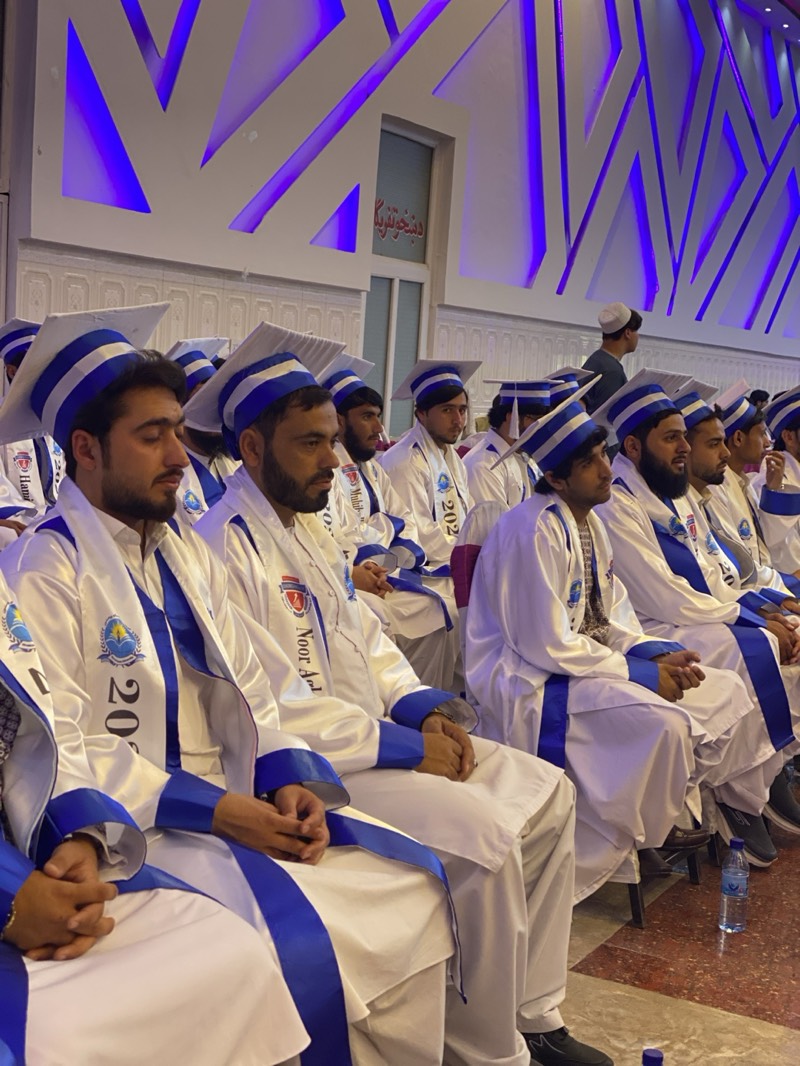 A medium, eye-level shot captures a group of young men seated in rows, presumably at a graduation ceremony, in Kandahar, Afghanistan. They are dressed in white gowns with blue trim and matching mortarboards. The gowns bear inscriptions, notably "2023" and "Noor Ahl," suggesting a graduation year and possibly the name of an institution. Most of the men have dark hair and beards.
The setting is indoors, marked by contemporary architecture featuring white walls and a decorative lighting design of blue angled lines on the upper walls. A door with signage in a non-English script, possibly Pashto or Dari, is visible. A plastic water bottle sits on the floor in the foreground. One man stands in the background wearing a white kufi hat. The overall atmosphere suggests a formal and celebratory event.