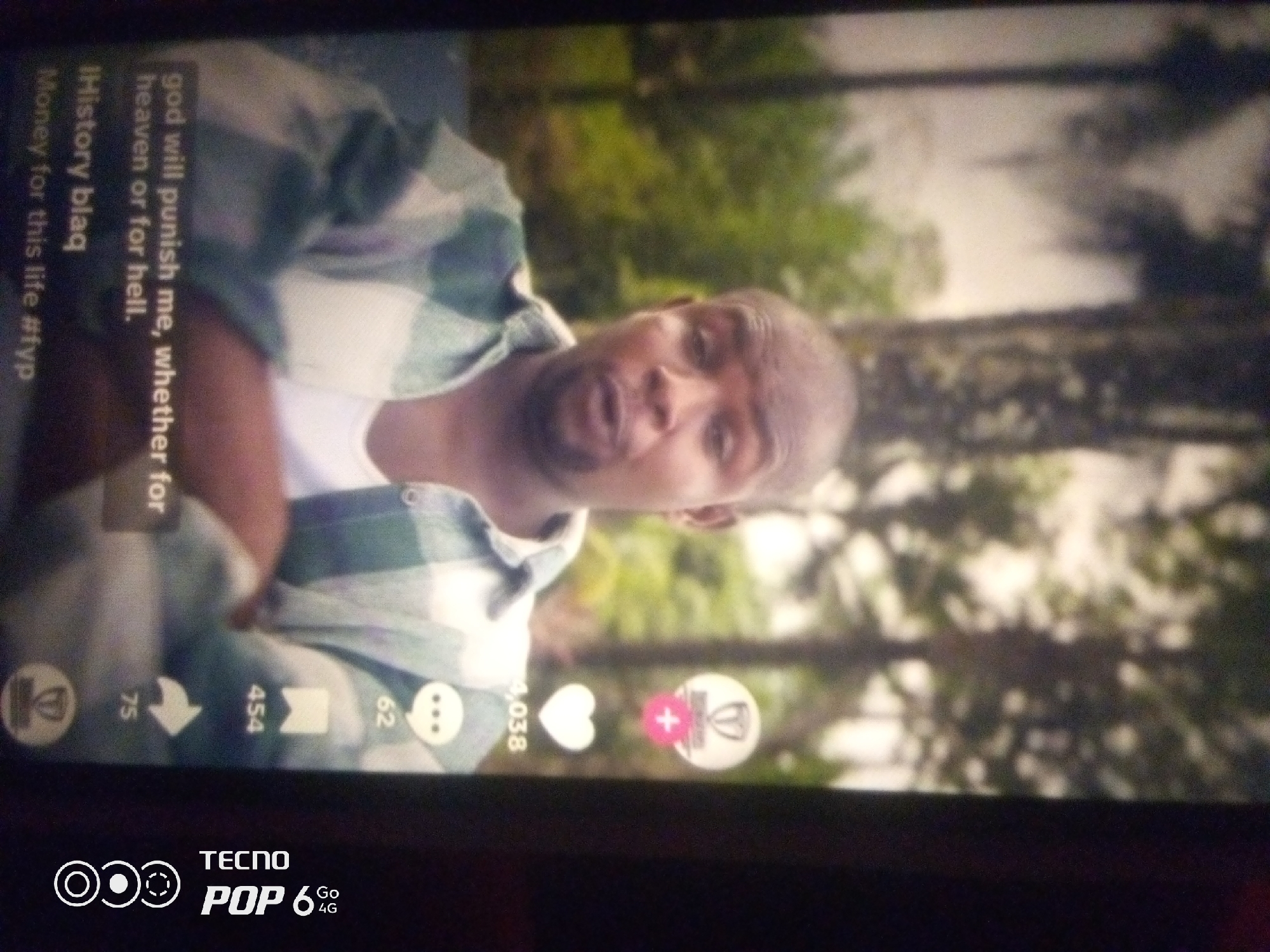 The image displays a close-up of a man's face and torso, captured from a low angle. He appears to be speaking, with his mouth open. He is wearing a green and white plaid shirt over a white undershirt. The background is blurred and consists of green foliage, suggesting an outdoor setting. Text overlays are visible on the left side of the image, including "god will punish me, whether for heaven or for hell.", "Money for this life #fyp", and "iHistory blaq". There are also icons and numbers at the bottom, typical of social media interfaces, indicating engagement metrics like "75", "454", "62", and "4,038" along with icons for likes, shares, and comments. A portion of a phone screen is visible at the bottom with "TECNO POP 6 Go 4G" branding. The image is taken in Howayi, Nigeria.
