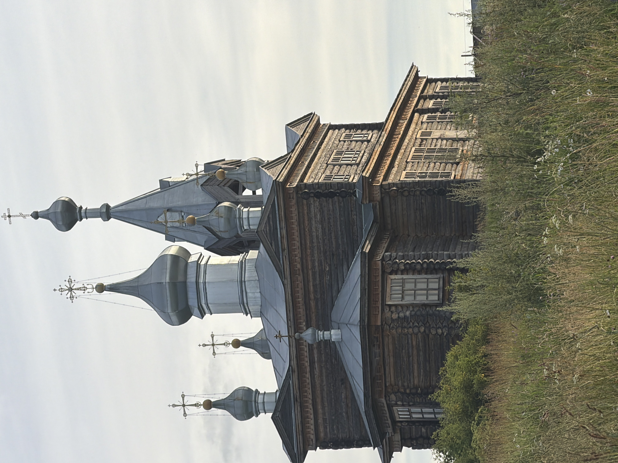 This is a vertical, eye-level, outdoor photograph of a traditional wooden Russian Orthodox church with multiple onion domes topped with crosses. The church's exterior is constructed from dark, weathered logs, and features several small, multi-paned windows. The domes are silver, and also topped with decorative crosses. The church is situated on a grassy hill with dense, dry vegetation and scattered wildflowers to the right. The sky is overcast and pale. The overall impression is one of rustic simplicity and age, likely taken in the daytime under diffused light. No people are visible in the image, and there is no visible text. The location is identified as Cherdyn, Russia.