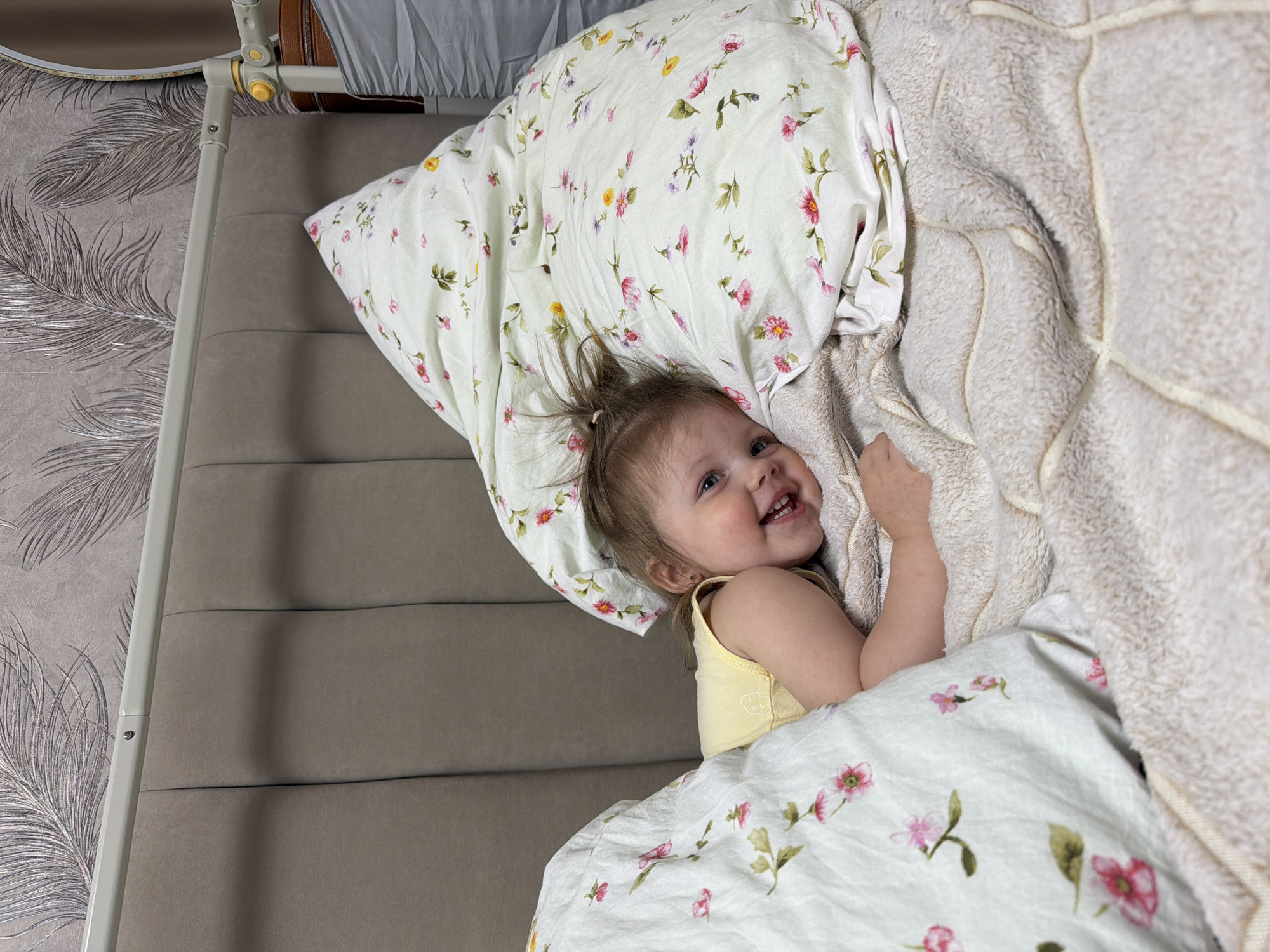 A young child is depicted lying in a bed in an indoor residential setting in Reshetnikovo, Russia. The child, with fair skin and light brown hair gathered in a small ponytail atop her head, is positioned on her left side, facing slightly towards the camera with a broad smile revealing her teeth. She has blue eyes and is wearing a light yellow sleeveless top with a small, indistinct light-colored graphic on the front. Her head rests on a large, white pillow covered in a floral-patterned pillowcase featuring scattered pink, yellow, and purple flowers with green leaves. A matching floral-patterned duvet or comforter partially covers her torso and lower body. Beneath the child and the duvet is a beige, textured, quilted blanket with distinct stitching patterns. The bed features a padded, upholstered headboard in a taupe or light brown fabric with vertical tufting. Above the headboard, a light-colored metal bar of the bed frame is visible, secured by a bolt. To the far right, a vertical section of the bed frame is visible, featuring a rounded, yellow adjustment knob. The wall behind the bed is covered with wallpaper displaying a repeating pattern of stylized metallic-grey feathers against a greyish-brown background.