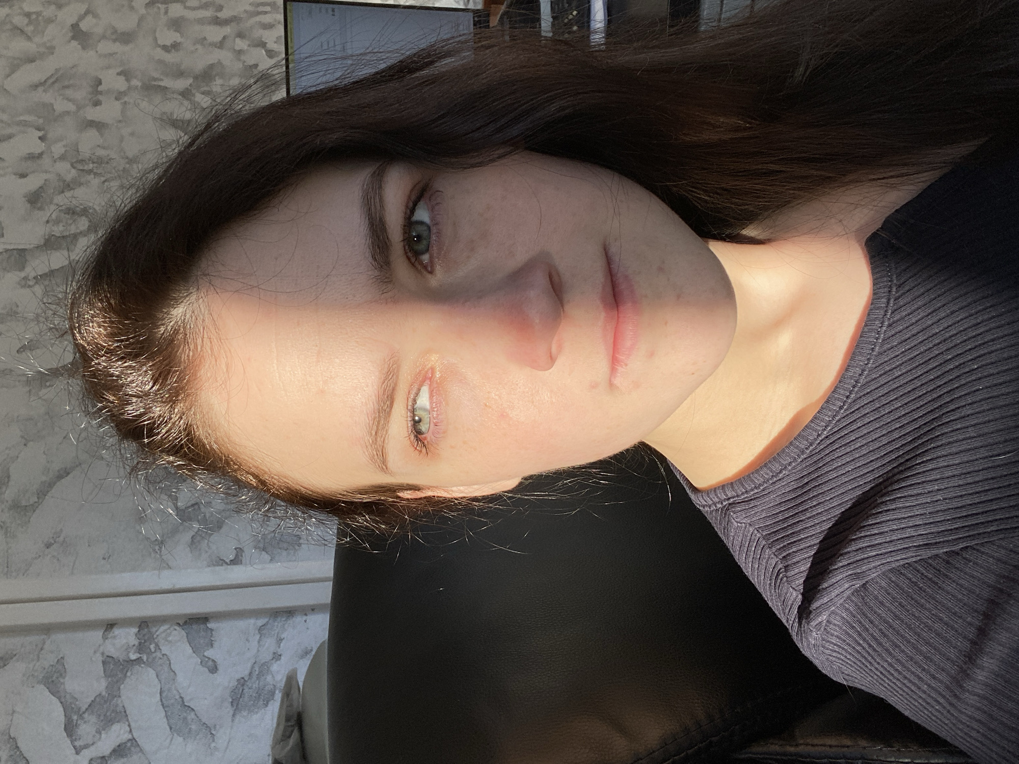 The image presents a close-up, rotated portrait of a young female taken indoors in Chaikovsky, Russia. She has long, dark brown hair, fair skin with noticeable freckles, and light-colored eyes, appearing greenish-grey. She is wearing a dark, ribbed top. The subject looks directly at the camera with a neutral and composed expression, her head slightly tilted.
The scene is characterized by strong, direct sunlight, which prominently illuminates the left side of her face (viewer's left), creating a distinct highlight and shadow effect. This suggests a bright daytime setting, possibly morning or late afternoon, with clear weather allowing sunbeams to enter.
The background features a light-colored wall with a textured, abstract greyish-white pattern, resembling painted brushstrokes or a unique wallpaper design. A dark, possibly leather, piece of furniture, like a chair or sofa, is partially visible behind her. The original image is oriented 90 degrees counter-clockwise.
Notable details include the fine hairs at her hairline, subtle skin imperfections, and a slight mark on her upper lip. No visible text is present in the image.