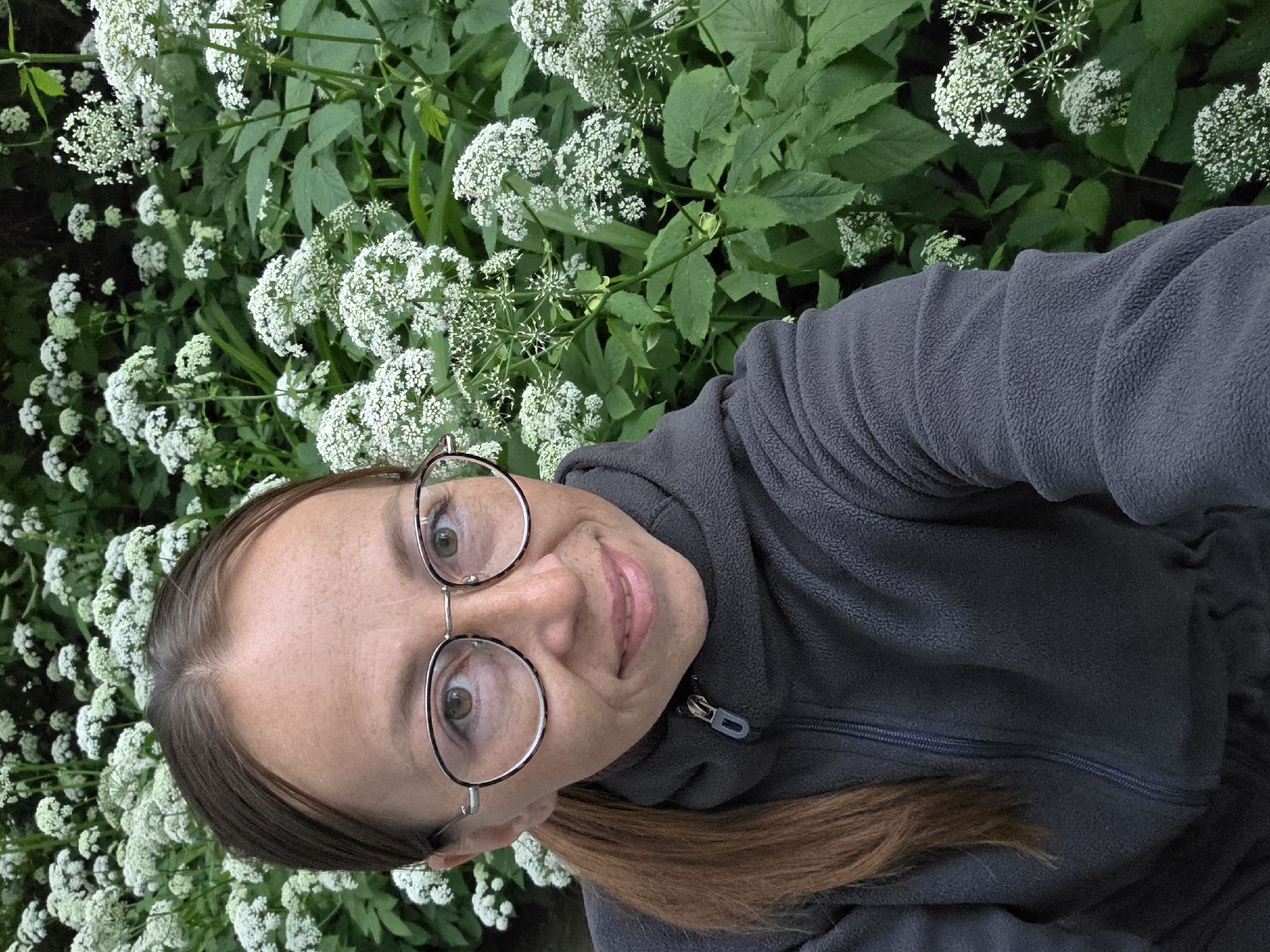 A female adult is depicted in a close-up self-portrait, centered slightly to the left, with an outdoor verdant background. The individual possesses light skin, brown eyes, and light brown hair parted in the middle, pulled back into a ponytail visible on the right side of the frame. She wears round metal-rimmed eyeglasses with a dark upper frame section. Her expression is a subtle smile with closed lips, and faint freckles are visible on her face. She is dressed in a dark gray fleece jacket with a high collar and a visible zipper extending down the front. The zipper pull is metallic with a dark blue or black accent. The background consists of dense green foliage interspersed with numerous clusters of small white flowers, characteristic of an umbellifer plant, suggesting a natural outdoor environment. The scene is located in the city of Chaikovsky, Russia.