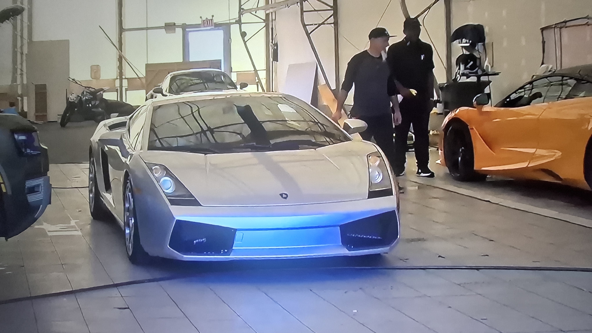 The image depicts the interior of a warehouse-style building. The space is occupied by several vehicles: a white Lamborghini with blue underglow lighting prominently in the foreground, an orange sports car on the right side, and at least two additional vehicles visible in the background. Two men are standing near the Lamborghini and the orange car; one is wearing a dark-colored shirt and a baseball cap, while the other is in all black. The background features an exit sign near a doorway, various pieces of equipment, and a motorcycle. The overall location appears to be a car customization or storage facility. The city and location is unknown.