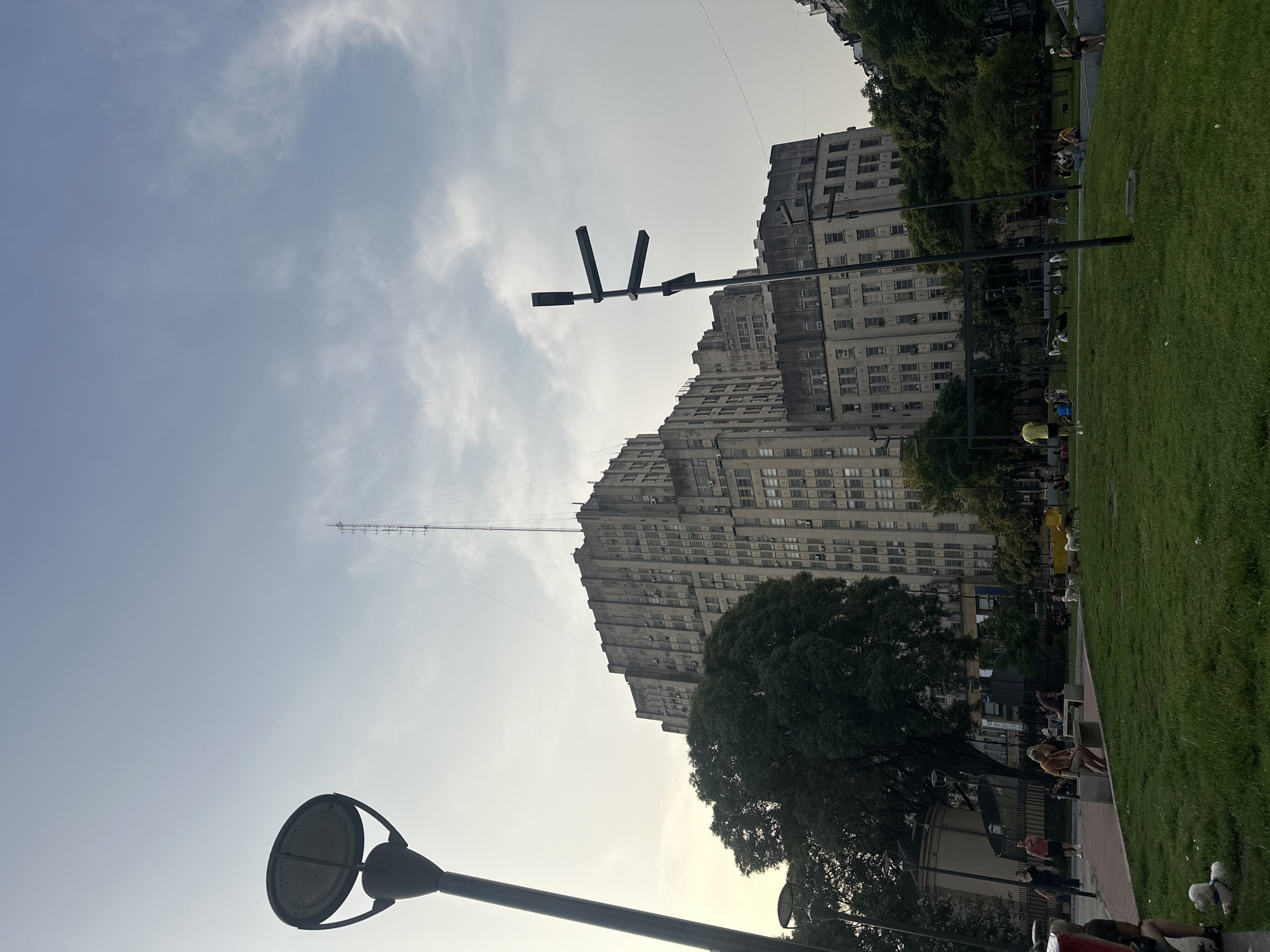 The image shows a scene in Buenos Aires, Argentina, specifically the Edificio del Ministerio de Desarrollo Social. The building is a large, multi-story structure with many windows, constructed of concrete. A tall antenna is affixed to the top of the building. In the foreground, there's a park area with green grass, trees, benches, and people. Some people are sitting on benches, others are standing or walking. Several modern-style lamp posts are visible in the scene. The sky is overcast with light clouds. The overall lighting suggests it's daytime, but not direct sunlight.