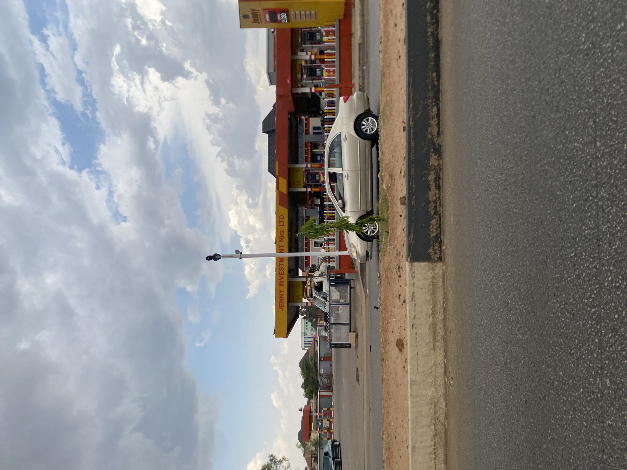 The image shows a Jenny Investment Nig. Ltd gas station in Gura Top, Nigeria. A light-colored sedan is parked adjacent to the station, alongside a road with a curb. The sky is cloudy. Other buildings and vehicles are visible in the background.