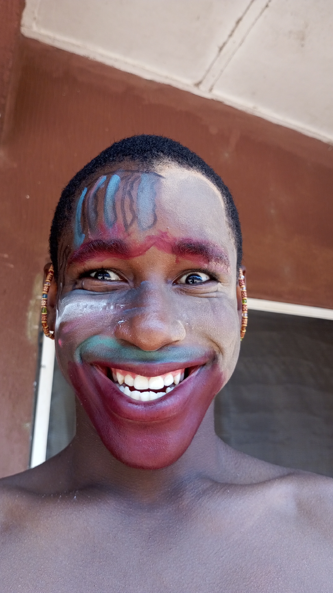 A close-up shot of a dark-skinned person with facial paint is taken in Nkpor, Nigeria. The person's forehead has a pattern of blue, grey, and red paint, while the eyebrows are accentuated with red paint. The lower part of the face, from below the nose to the chin, is painted with a wide, exaggerated red and green smile exposing white teeth. The person is wearing beaded earrings. The background features a red building and a white window frame. The subject appears to be shirtless.