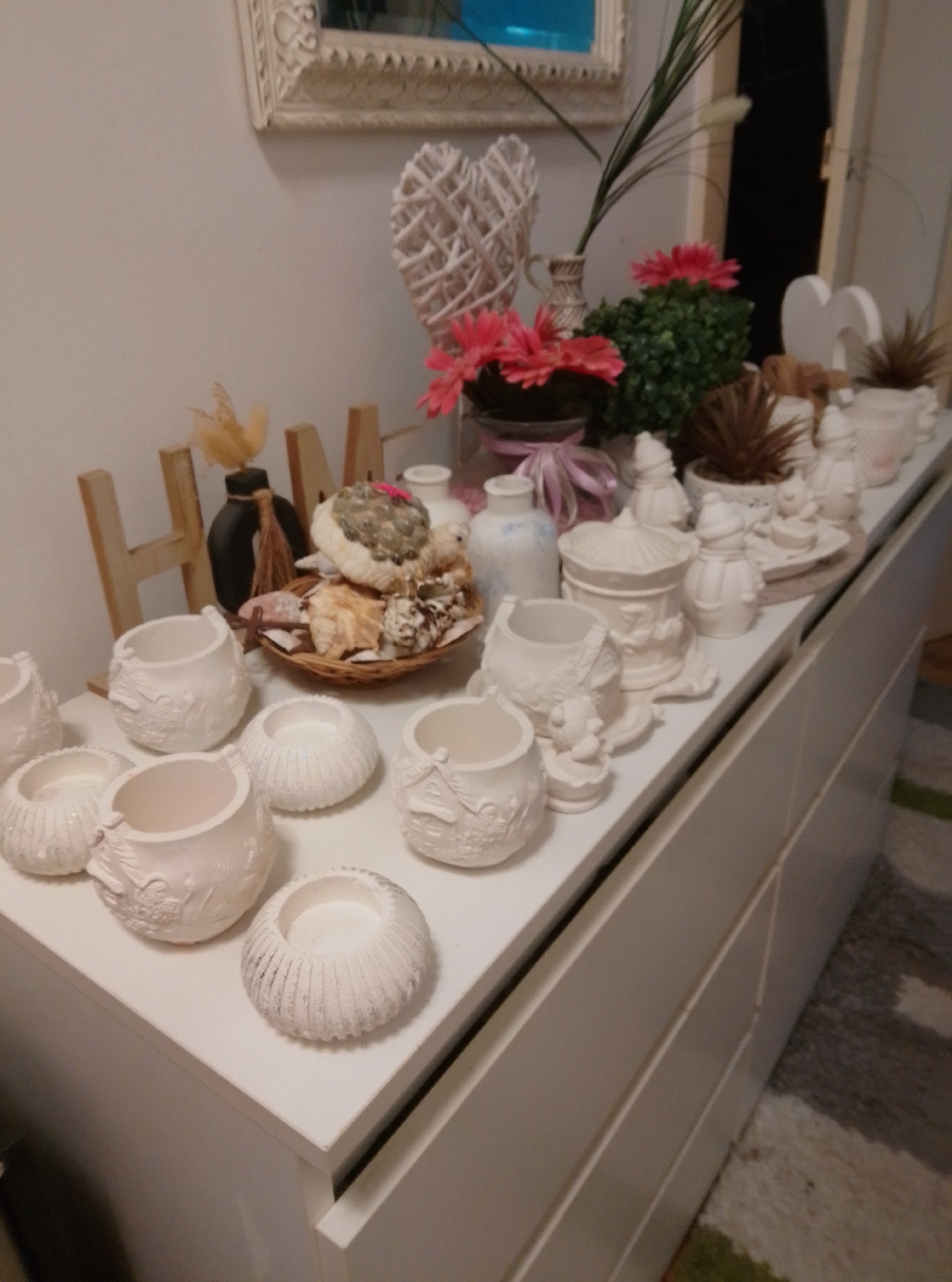 This is a high-angle, medium shot of a collection of white ceramic decorative objects displayed on a white dresser. The objects include various small pots, some with intricate embossed designs, and figurines. In the background, a woven heart decoration, artificial pink flowers, and a small potted plant are visible. The dresser is against a light-colored wall. The scene is indoors, and the specific location in the city of Wedel, Germany, cannot be determined from the image. There are no people or animals present in the image. The lighting is soft and diffused, suggesting an indoor environment.