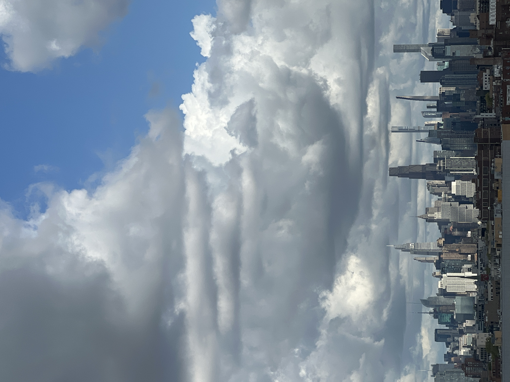 An expansive view captures the iconic skyline of New York City, specifically Manhattan, stretching horizontally beneath a dramatic, cloudy sky. The image is composed of a dense array of skyscrapers and urban buildings occupying the lower third of the frame, with the sky dominating the upper two-thirds.
The cityscape features numerous tall buildings, showcasing a mix of architectural styles, from classic art deco structures like the distinct spire of the Chrysler Building (visible towards the right of the skyline) to modern glass towers. The buildings are tightly packed, indicating the high density of the metropolitan area. Sunlight catches various facades, creating highlights and shadows that add depth to the urban fabric.
Above the city, the sky presents a dynamic weather scene. Large, textured clouds in varying shades of grey and white dominate. Patches of clear blue sky are visible, particularly in the upper left quadrant, offering a stark contrast to the heavy cloud cover elsewhere. Some clouds are bright and fluffy, while others appear denser and darker, suggesting a mix of light conditions and possibly changing weather. The overall lighting indicates daytime, likely mid-day or afternoon, with the sun illuminating parts of the clouds and buildings from an unseen angle.
There are no people or specific activities visible. No text is discernible in the image. The scene conveys the grandeur and intensity of a major urban center under a visually striking sky.