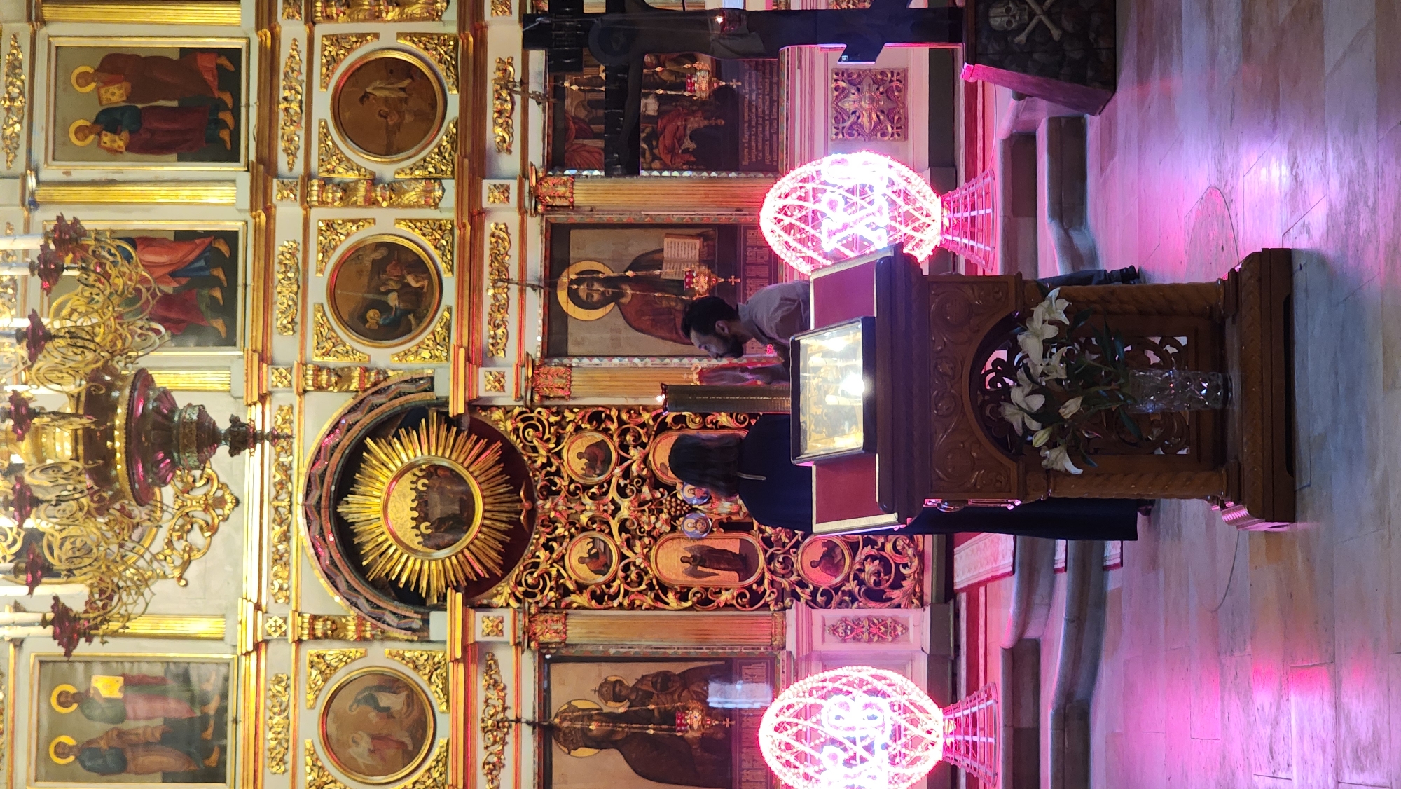 The image depicts the interior of an ornate Orthodox church, likely in Unknown, Unknown. A wall covered in religious icons and golden detailing is prominently visible. In the foreground, two individuals are bowing before a stand that holds a book. The stand is decorated with a vase of white lilies. Two round, illuminated pink light fixtures flank the stand. The floor appears to be marble. A large chandelier hangs on the left side of the image.