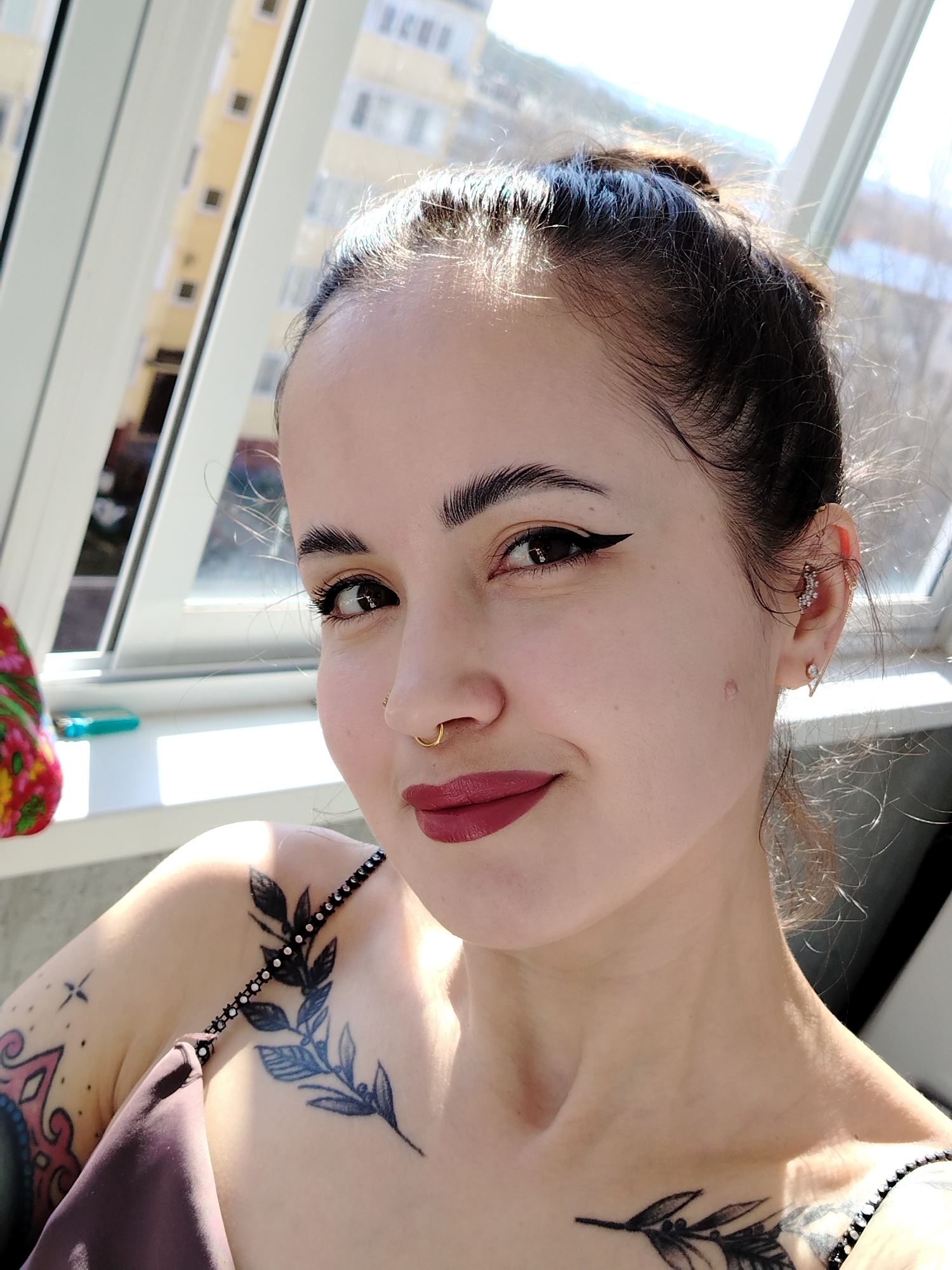 A medium shot captures a woman with dark hair styled in a bun, wearing a tank top. She has fair skin, dark eyebrows, and wears makeup, including eyeliner and dark lipstick. She has several piercings in her left ear and a nose ring. Tattoos are visible on her left shoulder and chest. The background shows a window with multiple panes and a building outside.