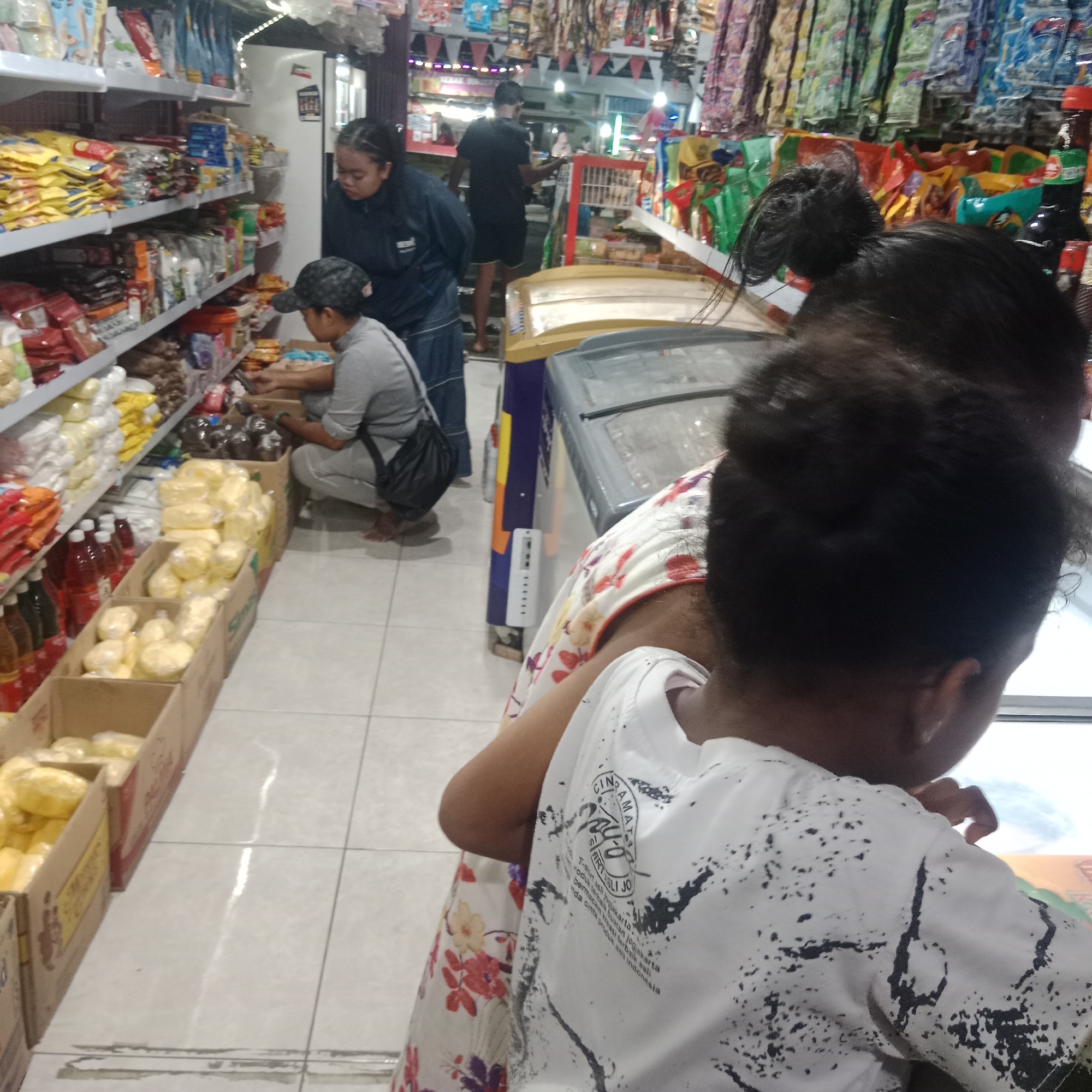 The image depicts the interior of a brightly lit convenience store, possibly located in Tegowanu Wetan, Indonesia. The aisles are stocked with various goods, including packaged snacks, drinks, and what appear to be fresh produce or dairy products in cardboard boxes. Several individuals are present in the store. In the foreground, a child wearing a white t-shirt with a black abstract pattern is visible from the back. To the left, an adult wearing a floral dress is partially visible. Further back, two other individuals are seen browsing the shelves. One individual, wearing a dark jacket and a baseball cap, is crouching down to look at items in a box. Another person, dressed in a dark blue jacket, stands near the shelves, possibly interacting with the individual in the cap. A man in a black t-shirt and shorts is visible in the background, near a red and white display unit. The store has tiled flooring and fluorescent lighting overhead, creating a well-lit environment.