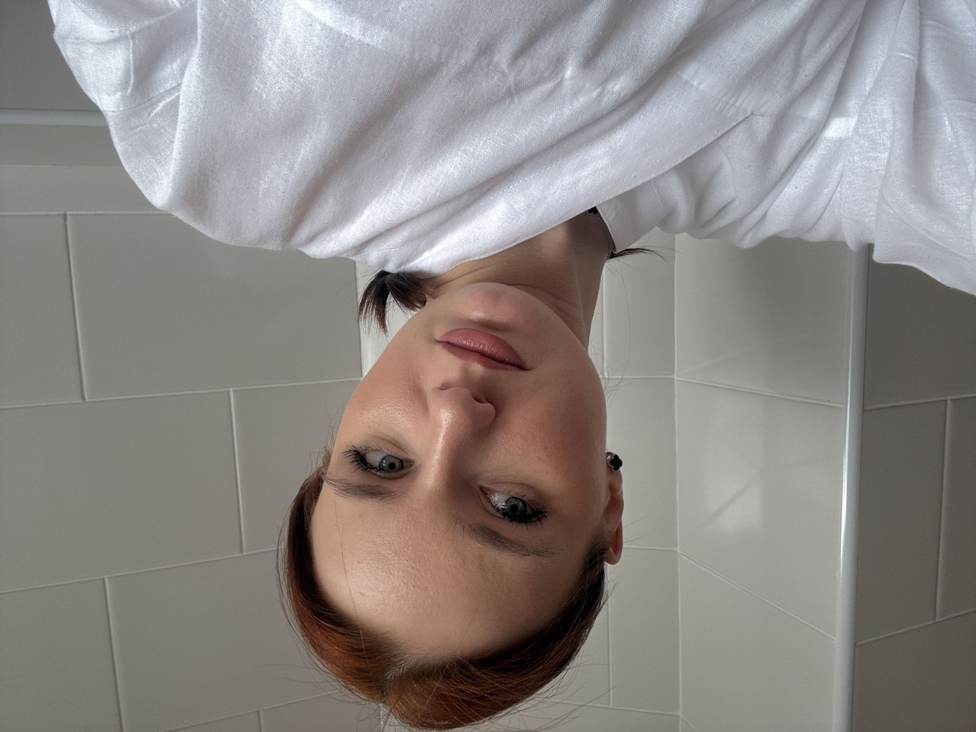 A Caucasian woman with red hair is looking down at the camera. She has light skin, blue eyes, and pink lips. She is wearing a white shirt or blouse. She is in a bathroom, with a tiled wall behind her. The location is not discernible from the provided image.