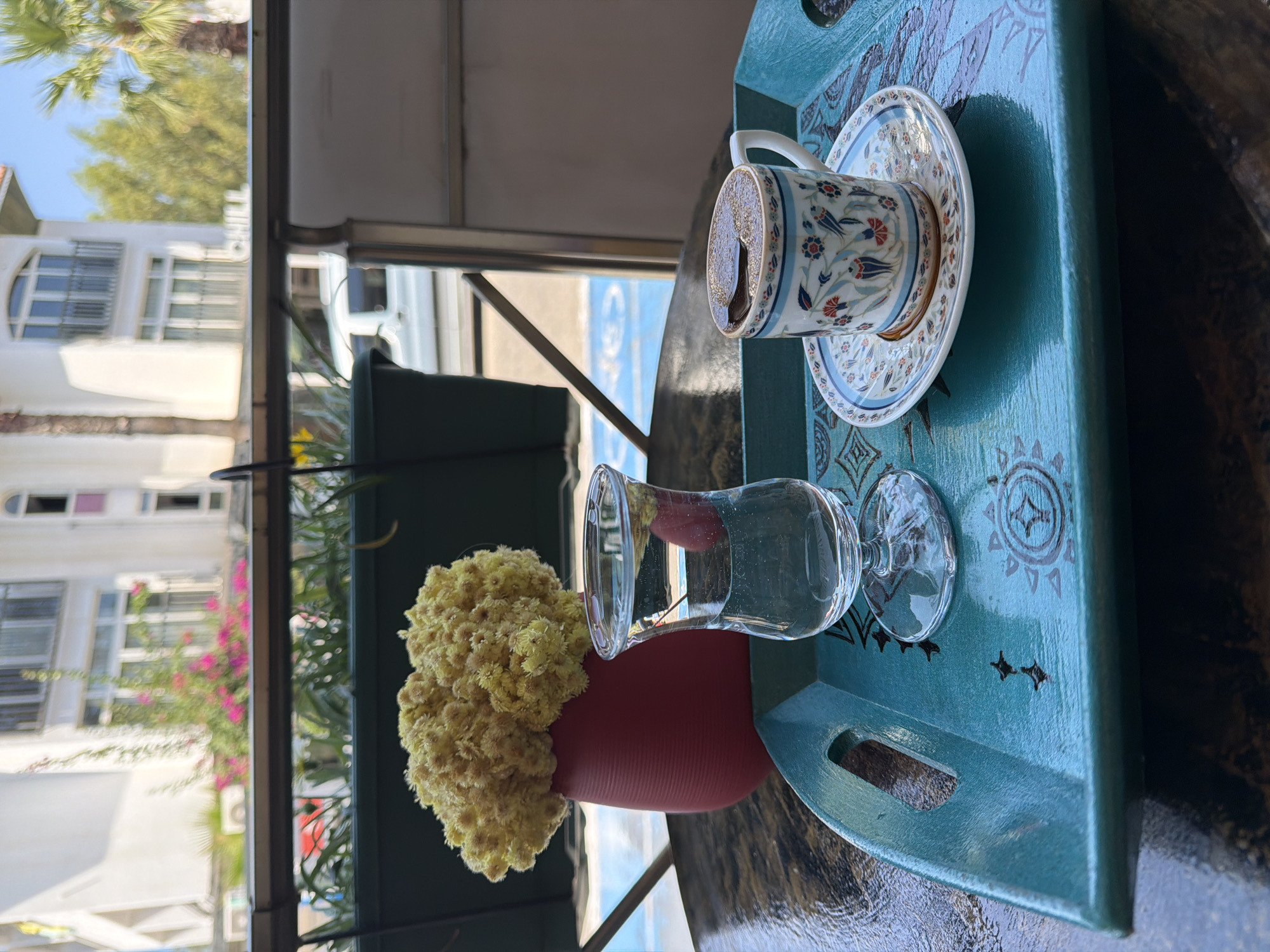 A teal-colored tray sits on a dark, patterned surface. On the tray, there is a small, decorative cup filled with Turkish coffee, a saucer with a floral design, and a clear glass of water. To the left of the glass is a red vase with a bouquet of dried yellow flowers. In the background, a balcony railing separates the tray from a blurred view of a white building with arched windows, green trees, and pink flowers. The scene appears to be outdoors, likely a cafe or balcony in Marmaris, Turkey, judging by the architecture and vegetation.
