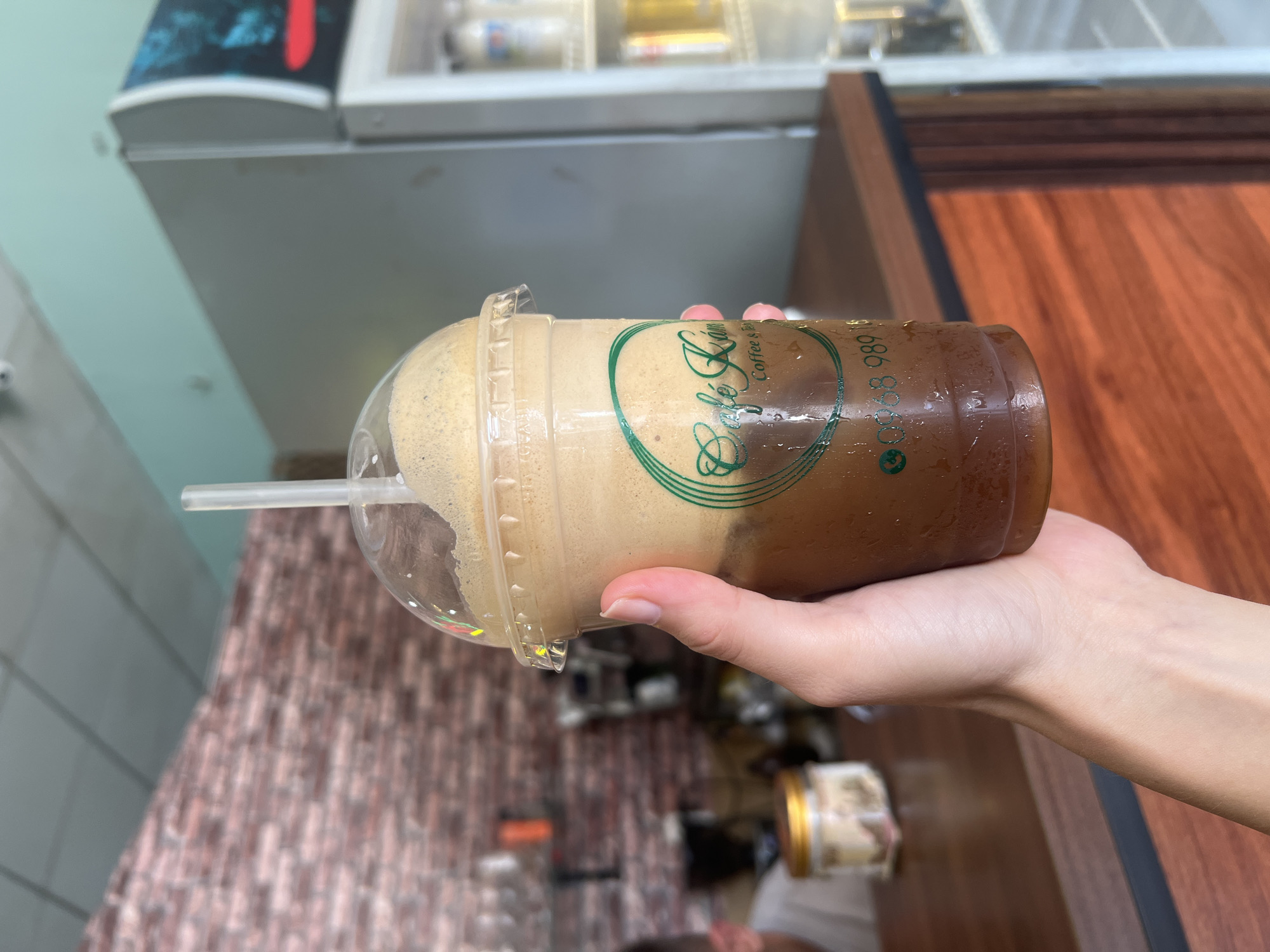 A person's hand holds a clear plastic cup filled with a layered cold beverage, likely an iced coffee, in what appears to be a cafe in Bắc Nha Trang, Vietnam. The drink features a distinct dark brown layer at the bottom, topped by a thick, lighter-colored foamy layer, typical of some Vietnamese coffee preparations. Condensation beads on the cup's exterior confirm it is a cold drink, and a clear plastic straw is inserted.
Visible text on the cup includes the cafe's name, "Cafe Kim," rendered in green script, with "Coffee & Tea" beneath it in a smaller font. Below this, a phone number "0906 583 989" is clearly displayed.
The background suggests an indoor setting. To the left is a light blue-green wall, while a white commercial refrigerator or cooler is partially visible in the upper background. A wooden counter or table occupies the right side of the frame. Below the held cup, a wall with a brick-like or patterned tile texture in shades of brown and pink is visible. Several blurred objects, including a jar with a golden lid, are present in the soft-focused background, hinting at a retail or beverage-selling establishment. The lighting suggests an indoor daytime scene.