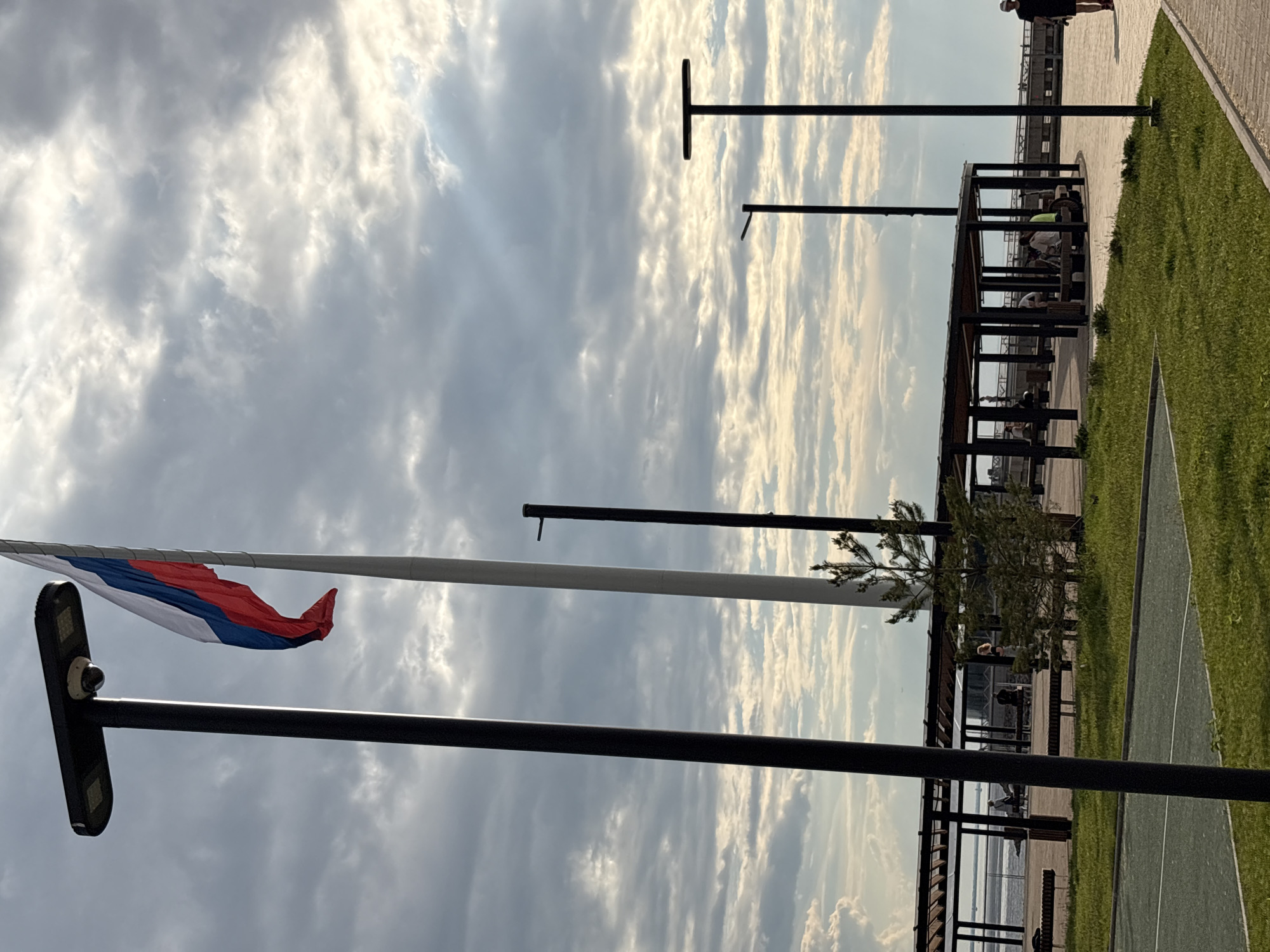 The image depicts an outdoor public space, likely a waterfront promenade or park, under a dynamic sky. The scene is illuminated by sunlight breaking through partially cloudy skies, suggesting late afternoon or early evening.
In the left portion of the image, a tall white flagpole proudly displays the Russian flag, with its white, blue, and red horizontal stripes gently unfurled. Below it, a modern, dark-colored lamppost features a flat, rectangular light fixture and what appears to be a surveillance camera or sensor on its upper side. Several other dark, slender poles, possibly streetlights or architectural elements, are visible throughout the foreground and midground.
The right side of the image showcases a structured recreational area. A long, open-sided pergola or shelter with a slatted roof provides shade over benches, where several people are seated or standing, engaged in leisurely activities. Adjacent to the pergola are paved walkways made of light-colored material, bordered by manicured green grass or turf that includes visible white markings, possibly for a recreational game. Small trees and shrubs are planted around the pergola.
In the background, beyond the park facilities, a wide body of water extends towards the horizon, indicating a riverside or coastal location. The overall atmosphere is calm and relaxed, with people enjoying the public amenities. Based on the visible flag and context provided, the location is Saint Petersburg, Russia. There is no legible text visible within the image.