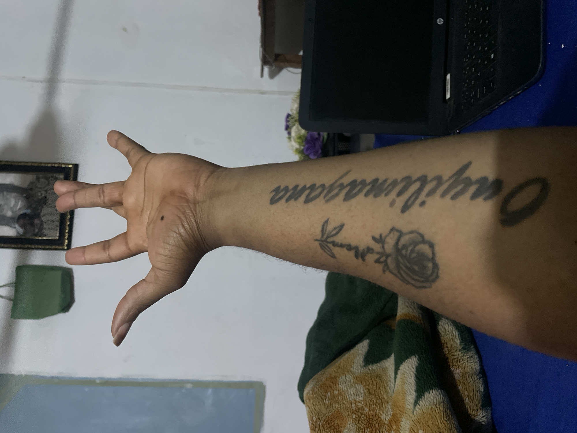 A forearm with a tattoo in Jos, Nigeria is visible. The tattoo includes the word "Onyilimagana" in a dark, sans-serif font and a rose. The hand is extended with fingers spread. A laptop and a framed photograph are in the background. Part of a blanket with a green and tan pattern is also visible. The background is mostly white walls.