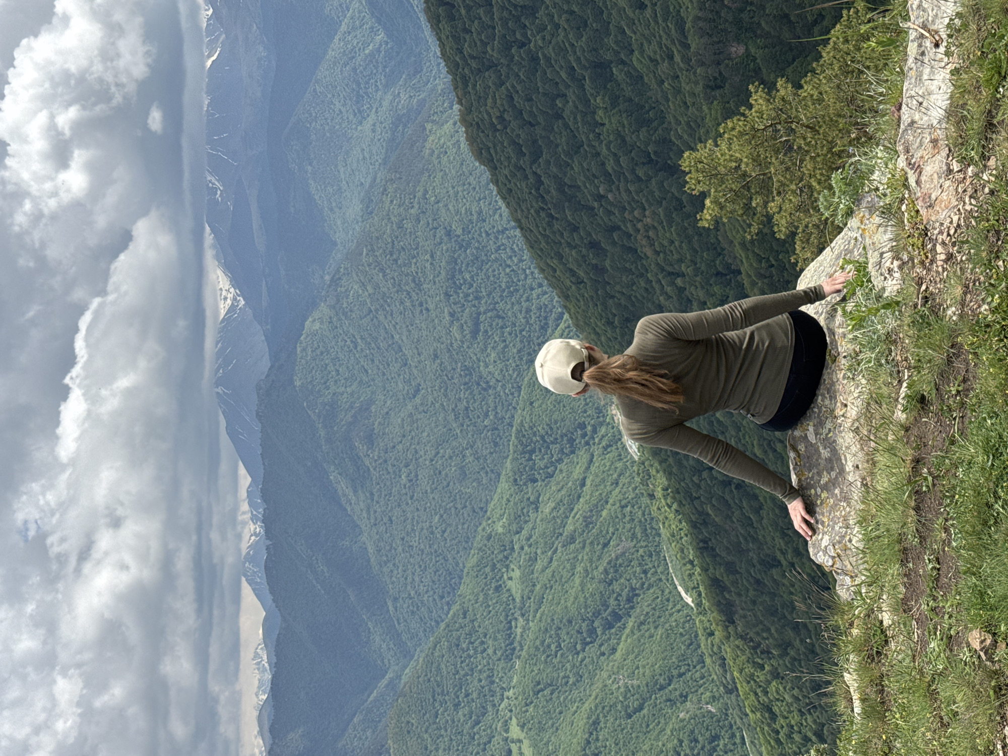 A woman wearing a green long-sleeved shirt, black pants, and a beige baseball cap sits on a rock overlooking a vast mountain landscape. The mountains are covered in dense green forests, with some snow-capped peaks visible in the distance. The sky is overcast with dramatic clouds. The location appears to be a scenic overlook in the mountains near Nizhnyaya Teberda, Russia. The woman is facing away from the camera, gazing at the panorama. The rock she is sitting on is large and irregular, with some patches of moss and lichen. The foreground consists of sparse green grass and some smaller rocks.