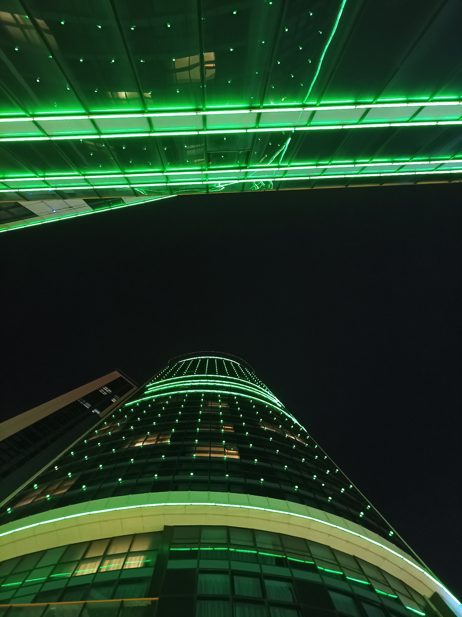 The image depicts a nighttime scene in Batumi, Georgia, featuring two prominent architectural structures illuminated with vibrant green lights. Above, a modern building or elevated walkway is seen with linear green neon lights spanning horizontally. Below, a taller, curved building, likely a hotel or office tower, is also outlined in green neon, with additional pinpoint lights covering its rounded facade. The surrounding environment is dark, suggesting a clear night sky above. The upward angle of the photograph emphasizes the height and modern design of the buildings.