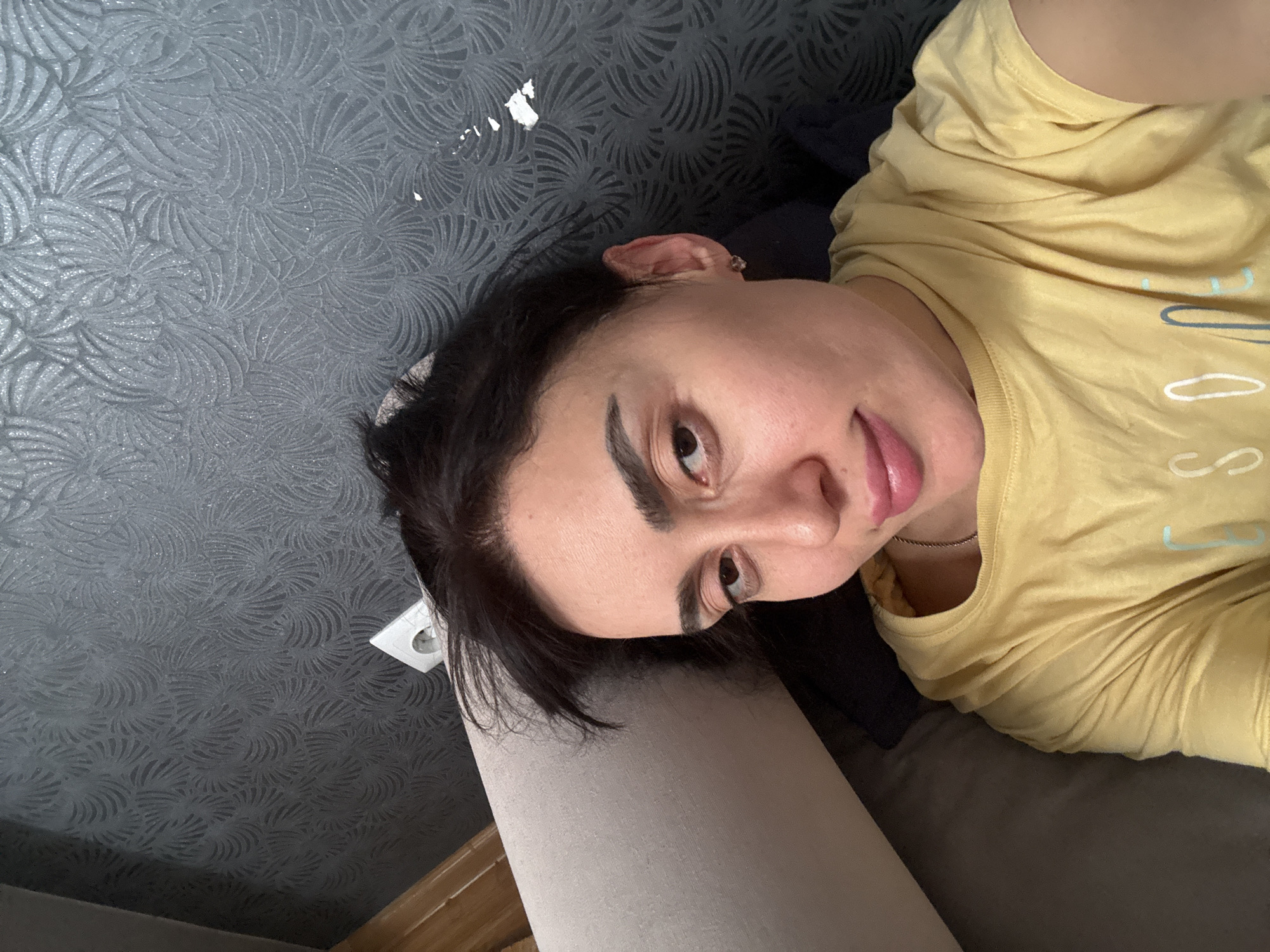 The image is a selfie of a woman lying on what appears to be a sofa or bed, with a patterned wallpaper in the background. The woman has dark hair and is wearing a yellow t-shirt. The location is not ascertainable from this image.