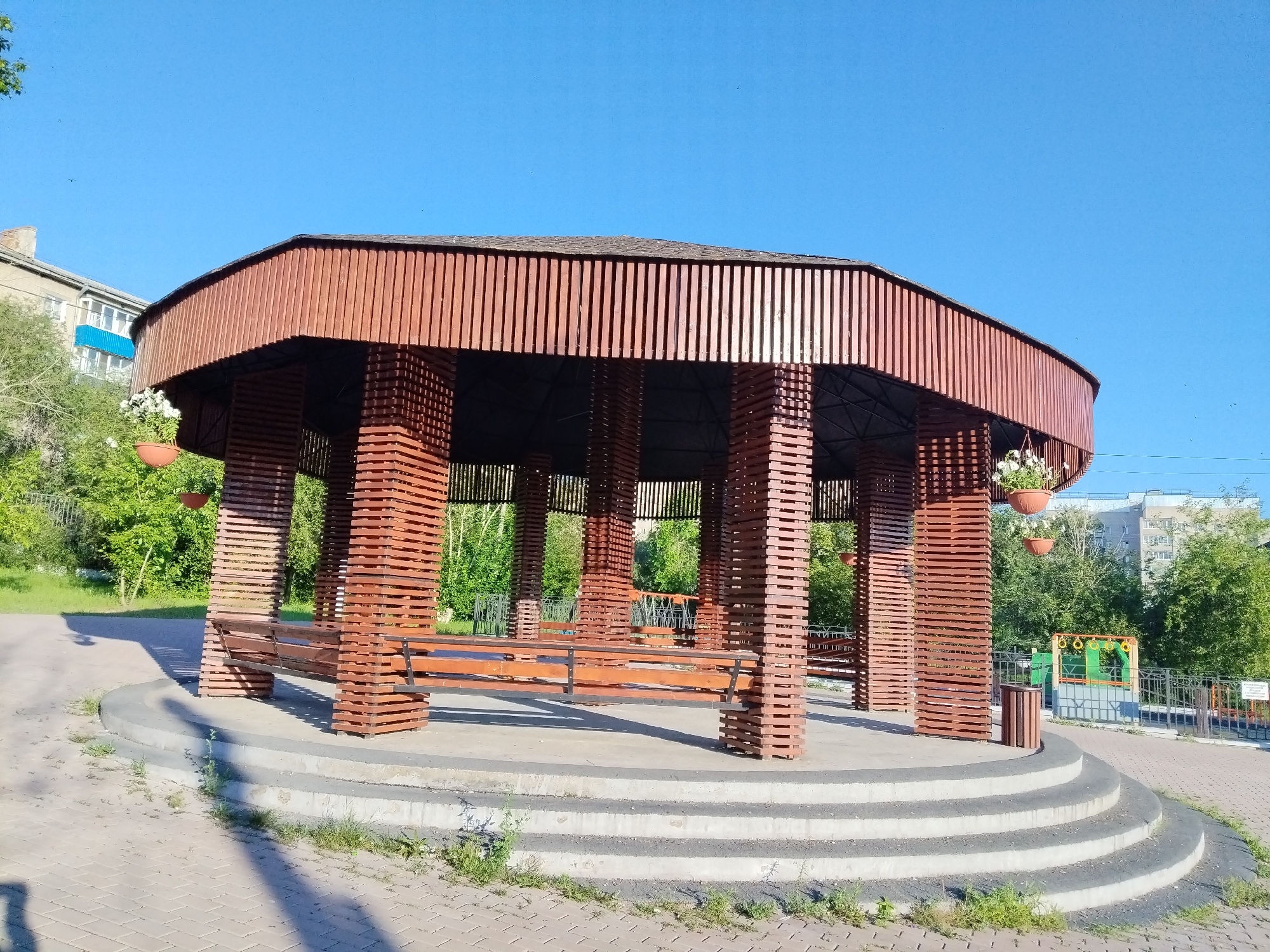 A wooden gazebo with benches inside stands on a concrete platform with tiered steps leading up to it. The gazebo has a rounded, octagonal roof supported by numerous vertical wooden slats that form the walls. Hanging planters with white flowers are attached to the exterior. To the left of the gazebo is a grassy area with trees, and behind it are residential buildings. To the right, a paved area leads to a playground with swings and a fence. The sky is clear blue, and the sun casts shadows. This scene is located in Chita, Russia.