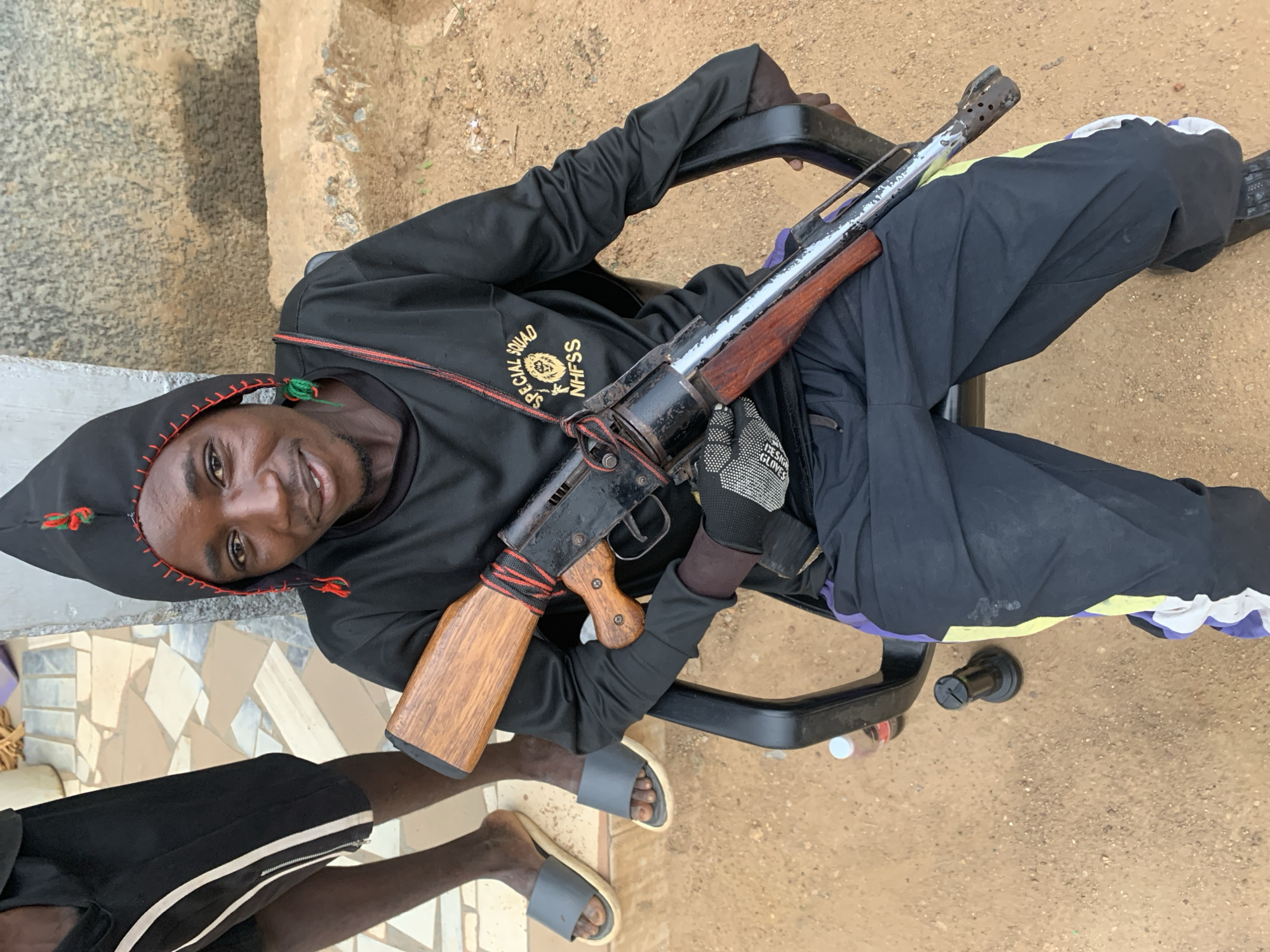 A person in a black hooded sweatshirt with the words "SPECIAL SQUAD NHFSS" is sitting in an office chair holding a rifle. The person has a black hood with red trim and green tassels, and a glove on the right hand. The person is looking at the camera and smiling. The chair is on a dirt ground next to a tiled surface where another person is standing. The location is New Stadium, Nigeria.