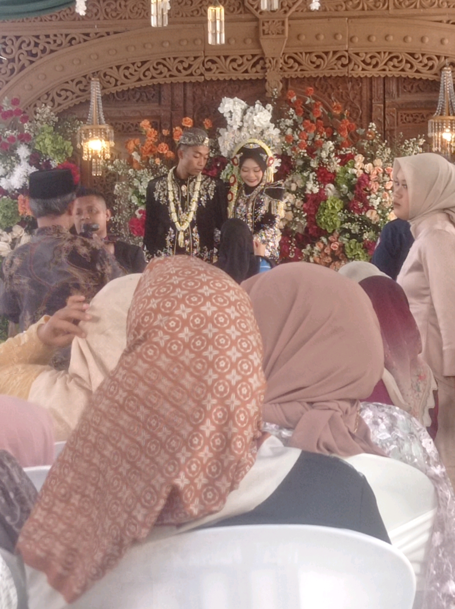 The image depicts a wedding ceremony in Ngemplak, Indonesia. In the background, a couple in traditional attire stands before a floral arrangement and ornate wooden decor. The groom wears a black outfit with a beaded necklace, and the bride is adorned with a traditional headdress and embroidered clothing. Several guests are present, some of whom are partially visible in the foreground, including individuals wearing headscarves and traditional clothing. The setting appears to be indoors, with decorative lighting fixtures hanging from the ceiling. The scene captures a moment of celebration and cultural tradition.