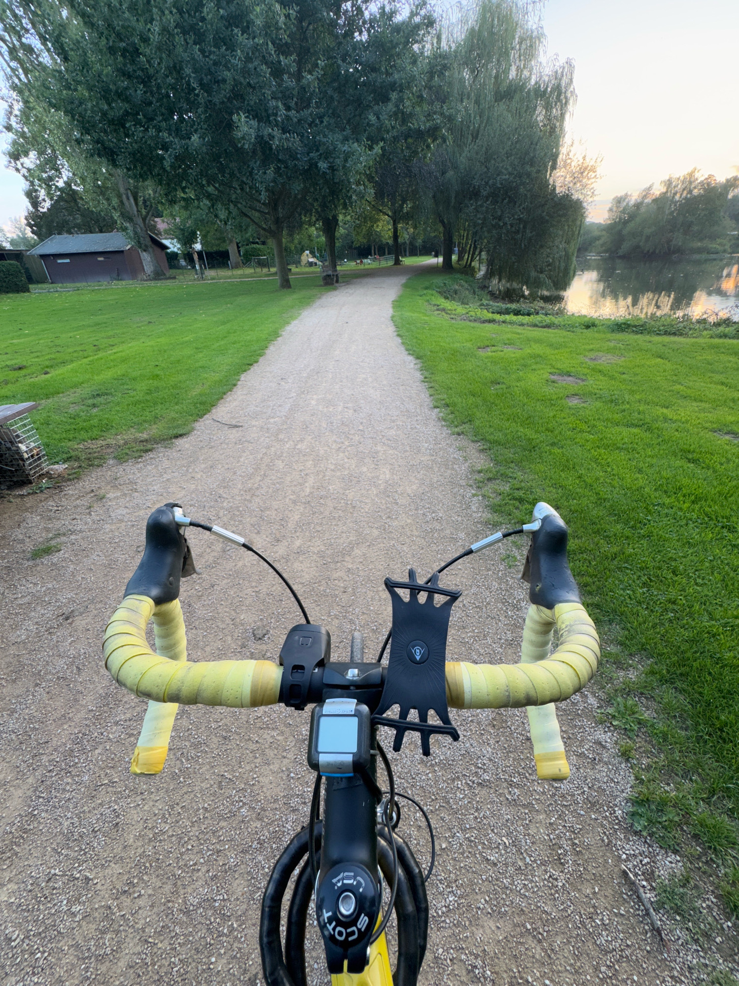 The image is a first-person perspective of a bicycle's handlebars on a gravel path. The handlebars are wrapped in yellow tape, and a phone mount is attached. A cycling computer is also visible on the handlebars. The path is lined with green grass and trees. In the background, a body of water, possibly the Rhine River, is visible. The sky is overcast, suggesting either early morning or late evening. The location is identified as Cleves, Germany.