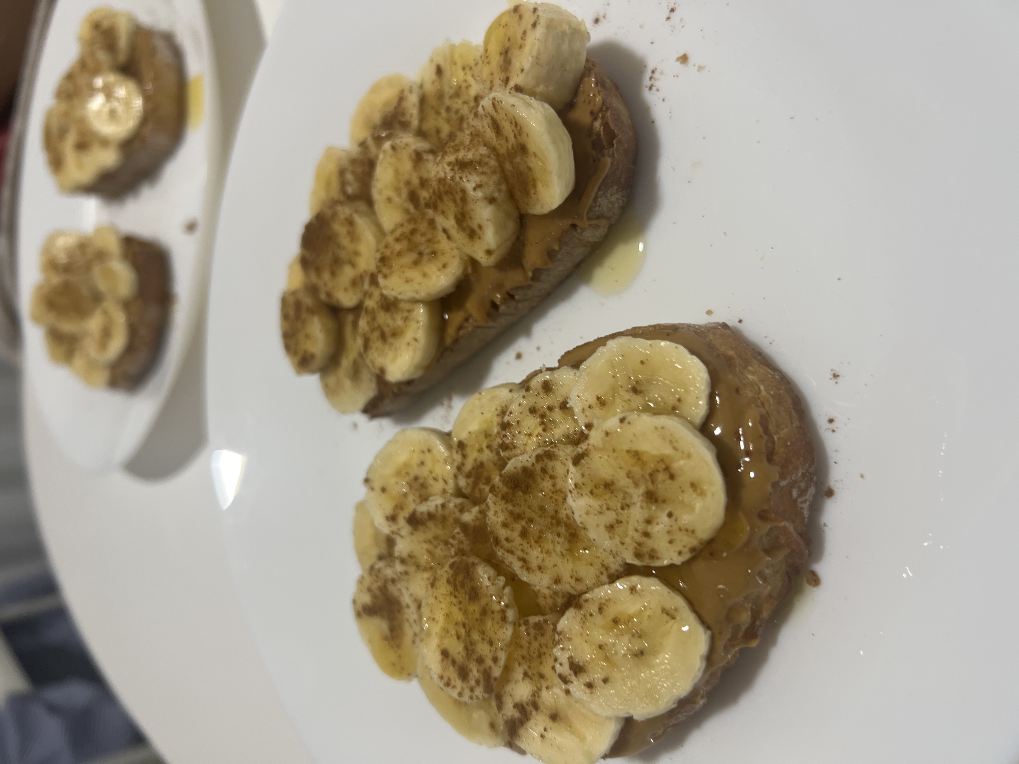 The image shows a plate of banana toast with a sprinkle of cinnamon. There are two pieces of toast in the center of the frame and two pieces of toast on a plate in the back left of the frame. The toast is arranged on a white plate. The image shows a close-up view of the food. The location of the image is not mentioned.