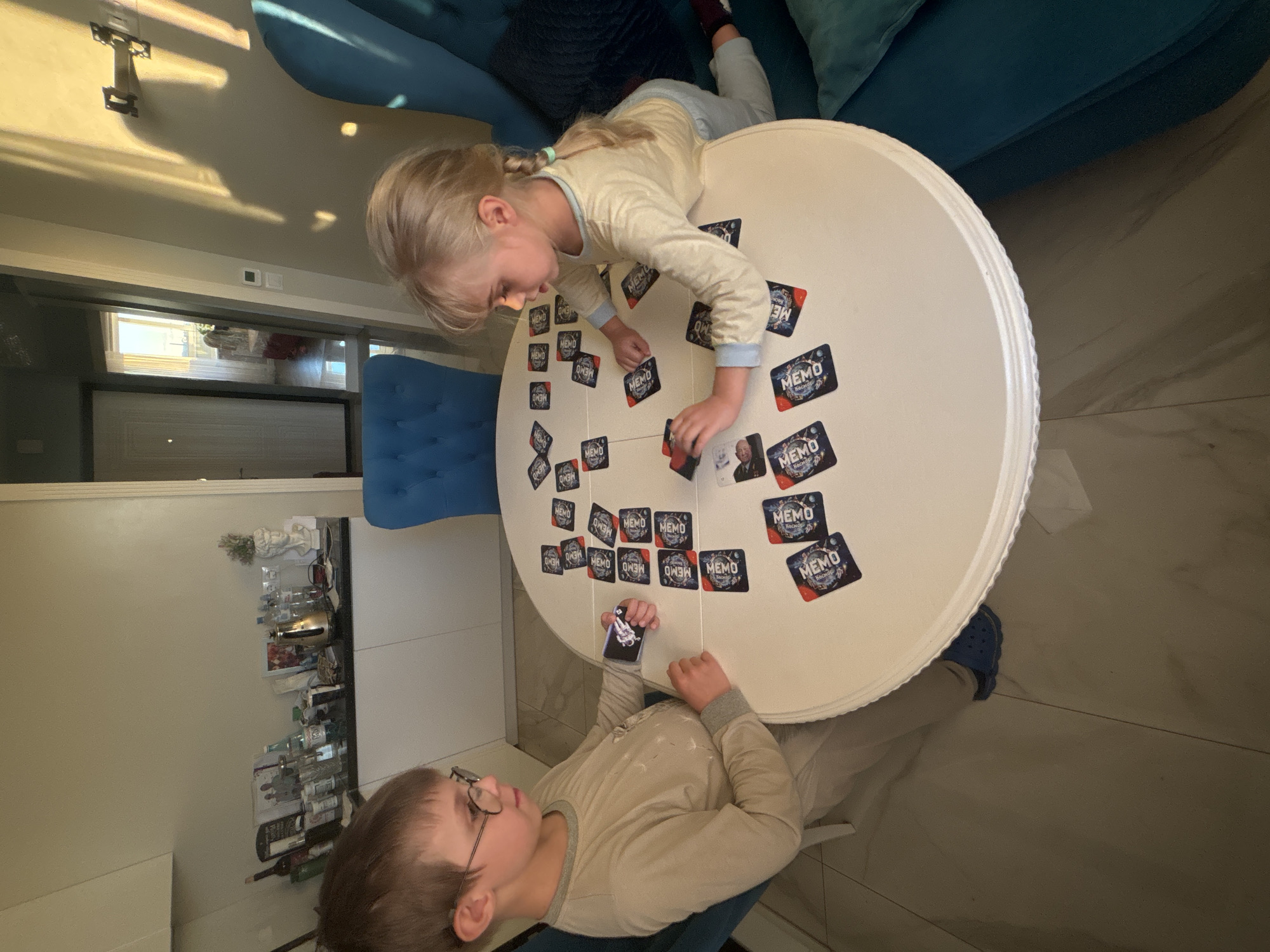 Two children are playing a memory card game at a round white table. A girl with blonde pigtails is leaning over the table, reaching for a card. A boy with glasses is sitting opposite her, looking at the cards. Many cards with the "MEMO" logo are spread face down on the table. The scene appears to be indoors, with sunlight streaming in from a window, casting shadows on the ceiling. A blue sofa is visible in the background, and a blue chair is positioned near the table. A shelf with bottles and decorative items is also visible in the background. The children seem engaged in the game.