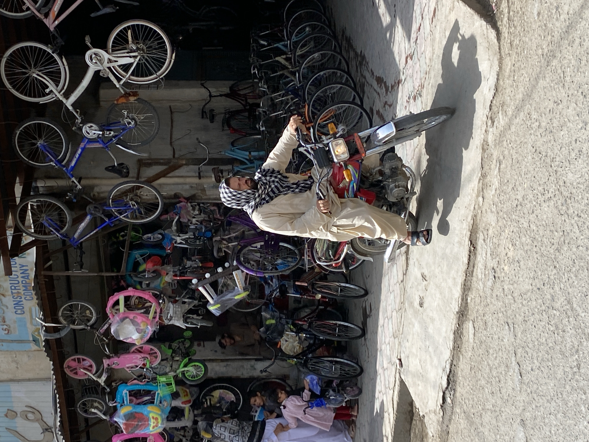 The image captures a street scene in Kandahar, Afghanistan, likely outside a bicycle shop. The foreground is dominated by a man riding a red motorcycle. He is wearing a light-colored robe and a black and white checkered headscarf. The motorcycle has a visible engine, handlebars, a front wheel with spokes, and a headlight. The man's right hand is on the handlebars, and his left hand is not visible. He is looking forward, seemingly engaged in riding.
Behind the motorcycle, a multitude of bicycles are displayed. Some are hanging from the roof of an awning, while others are lined up on the ground. These include children's bicycles in bright colors like pink and blue, as well as adult-sized bicycles in various states of repair and style. Many of the bicycles are suspended by their wheels or frames.
To the left of the motorcycle, a group of people are gathered. A woman in a white and patterned garment stands with her hand on the shoulder of a young girl wearing a pink dress and red leggings. The girl holds a blue umbrella. Another young child, a boy, stands nearby. Further back, within the shop entrance, a man with a beard and dark clothing sits.
The environment appears to be an outdoor marketplace or commercial area. The ground is paved with concrete and some sections show wear and tear. The background consists of a building with a corrugated metal roof and a sign with Arabic script and the words "CONSTRUCTION COMPANY". The overall lighting suggests it is daytime, with shadows cast on the ground, indicating the sun's position. The scene conveys a sense of everyday life and commerce in the city.