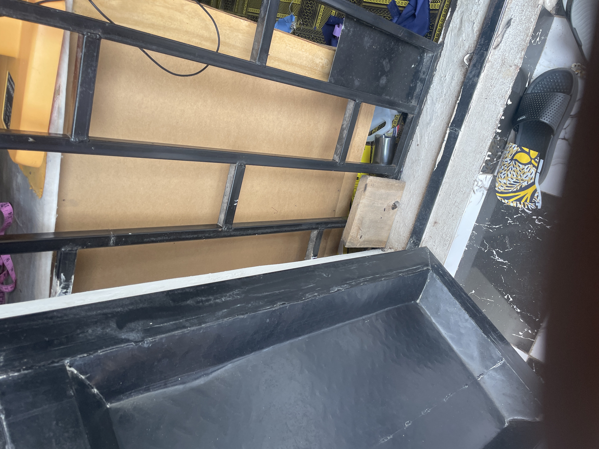 The image shows a close-up, overhead view of a black plastic basin, likely a laundry basin, placed on a patterned tiled floor. To the left of the basin, a black metal frame is visible, supporting a brown board. A yellow object and a measuring tape are partially visible beneath the frame. Further to the left, a portion of a yellow plastic container with some text is visible. To the right of the metal frame, a black flip-flop is placed on the tiled floor, with a piece of fabric featuring a yellow and black pattern underneath it. In the background, behind the metal frame, there is a glimpse of a yellow and black patterned fabric. The scene appears to be set indoors or in a covered outdoor area. The lighting suggests it is daytime. There is no clear indication of specific activities or emotions.