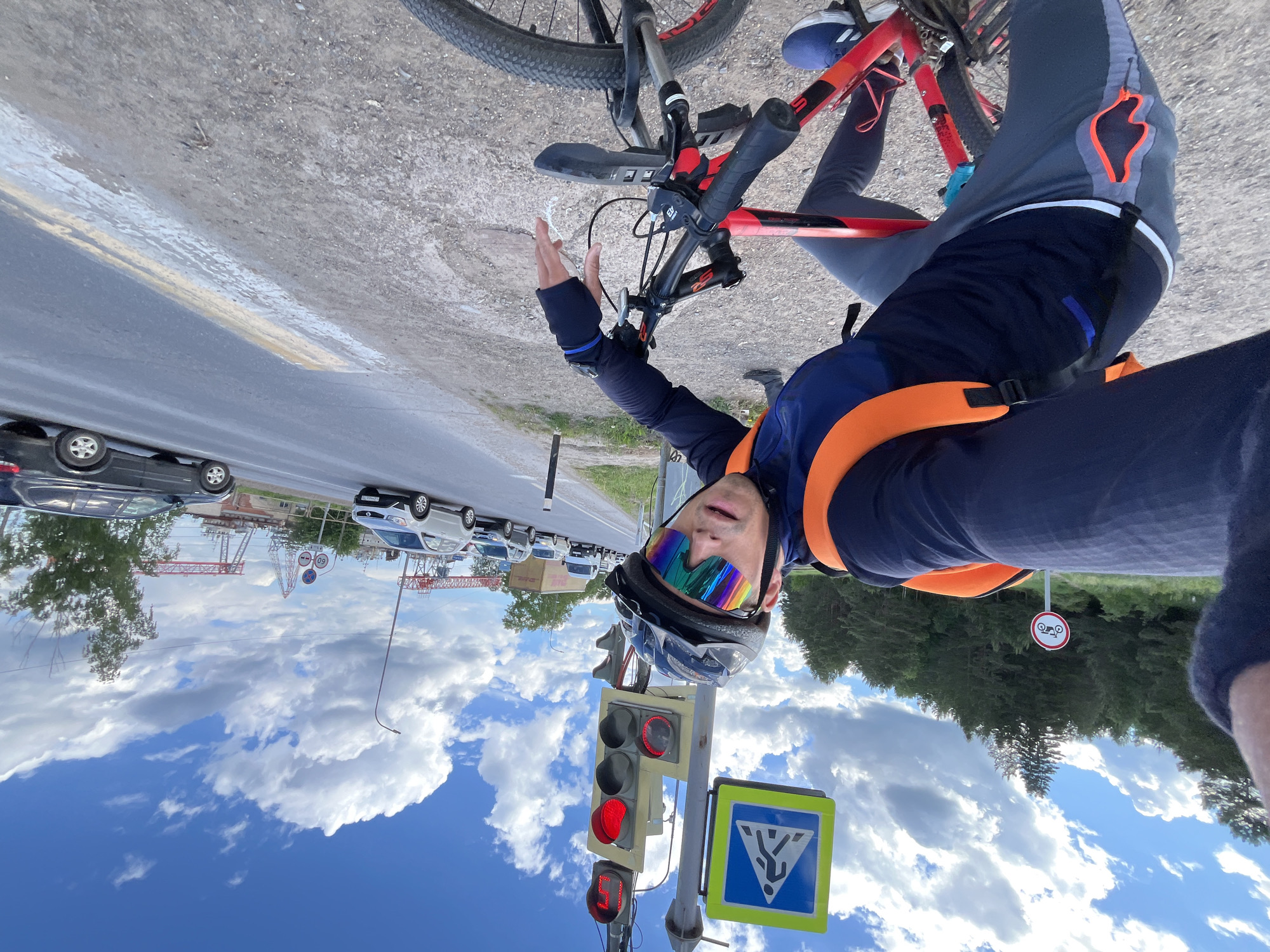 A man is riding a red and black mountain bike on a road in Chaikovsky, Russia. The traffic light is red, and a pedestrian crossing sign is visible. Behind him is a busy street with cars, and construction cranes are visible in the background. The sky is blue with white clouds. He is wearing a helmet and colorful sunglasses. A sign with a motorcycle and the number 50 is visible in the background. He is holding one arm up.