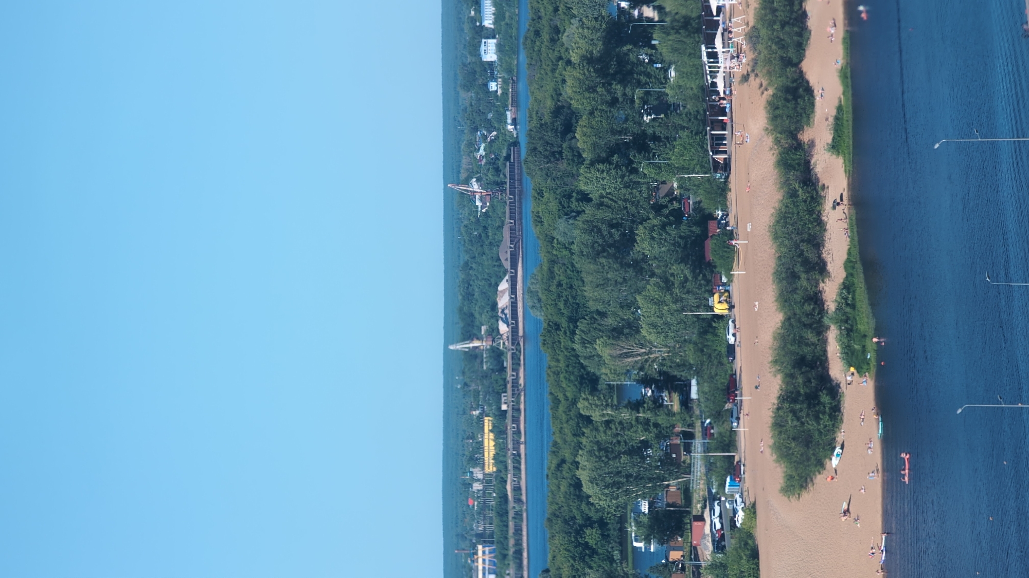 This aerial photograph depicts a sun-drenched day at a beach and riverfront, likely in Nizhny Novgorod, Russia. The scene is divided into three distinct zones: a clear blue sky occupies the left third of the frame, transitioning to a lush green expanse of trees and buildings along the riverbank, and finally, a sandy beach and a calm blue river.
On the beach, numerous people are engaged in various leisure activities. Some are sunbathing, others are strolling along the shore, and a few are swimming or wading in the river. Beach umbrellas and lounge chairs dot the sandy area, suggesting a popular recreational spot. A yellow inflatable boat is visible near the water's edge, adding to the recreational atmosphere. Along the tree-lined embankment, there are indications of a town or resort, with some buildings and parked cars.
The river itself is wide and relatively calm, with a few small ripples on its surface. Several lampposts are strategically placed along the riverbank, suggesting this area is well-maintained for public use, possibly a promenade or park. In the distance, industrial elements are visible, including a large crane and what appears to be a loading dock or port facility, hinting at the city's economic activity. A small plane is also visible in the sky, adding a dynamic element to the scene.
The bright sunlight and clear sky indicate a pleasant, warm weather day, likely in the late morning or early afternoon. The overall mood is relaxed and leisurely, characteristic of a summer day spent outdoors by the water. No visible text is present in the image.