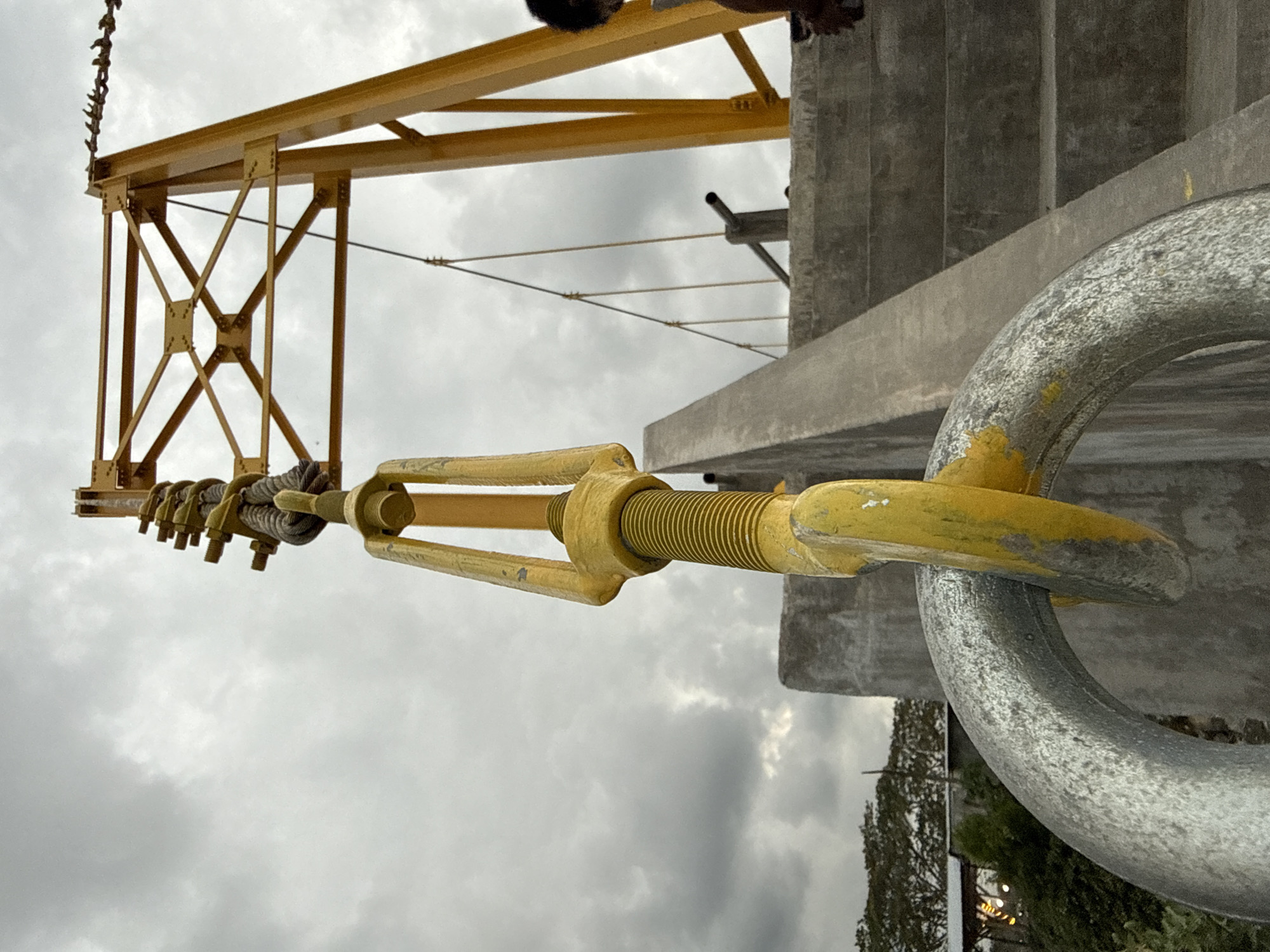 A low-angle shot shows a portion of a yellow metal construction crane with a long, yellow turnbuckle attached to a large gray metal shackle. The crane's structure features a lattice-like design with bolted intersections. The turnbuckle connects to the crane via a series of rope knots and metal loops. The shackle is connected to a gray concrete structure. Overcast skies filled with clouds are visible in the background. Partial views of human figures appear at the top of the frame. Vegetation, including trees and possibly distant buildings with lights, is visible in the lower right corner. No specific details confirm the location as Lhokseumawe, Indonesia.