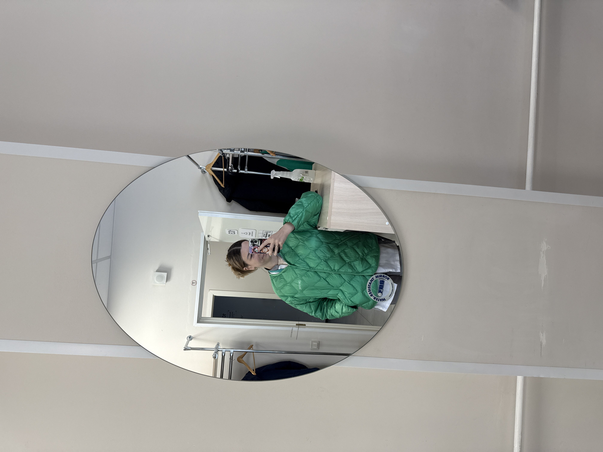 The image shows a mirror reflection of a person wearing a green quilted jacket, taking a selfie with a smartphone. The reflection is captured in an oval mirror mounted on a light beige wall. The background features a clothing rack with hanging garments, a doorway, and a counter. A sticker labeled "WATER-RESISTANT MIRROR FBS" is visible on the mirror's surface. The location may be a retail store or a dressing room. The image provides no specific information indicating the location is in Moscow, Russia.