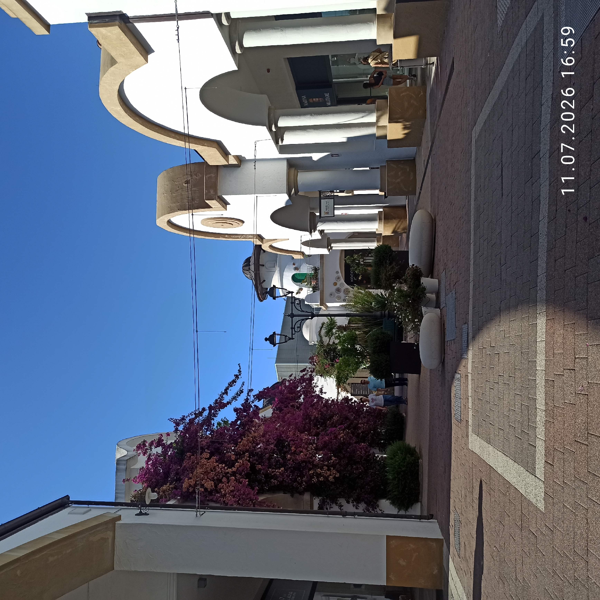 The image depicts a pedestrian walkway lined with white buildings featuring arched entrances and columns. A large, vibrant bougainvillea plant with deep purple and pink flowers cascades down the side of one building on the left. In the background, a distinct white tower with a dark domed roof and a green door is visible, characteristic of the architecture found in Molfetta, Italy, potentially within a shopping or marina area. The walkway is paved with reddish-brown bricks and has inlaid white lines. Several planters with greenery and decorative stone benches are placed along the walkway. Two individuals are walking away from the camera in the middle ground, and two women are visible in the far right background near a storefront. The sky is clear and blue. A timestamp "11.07.2026 16:59" is overlaid on the lower right portion of the image.
