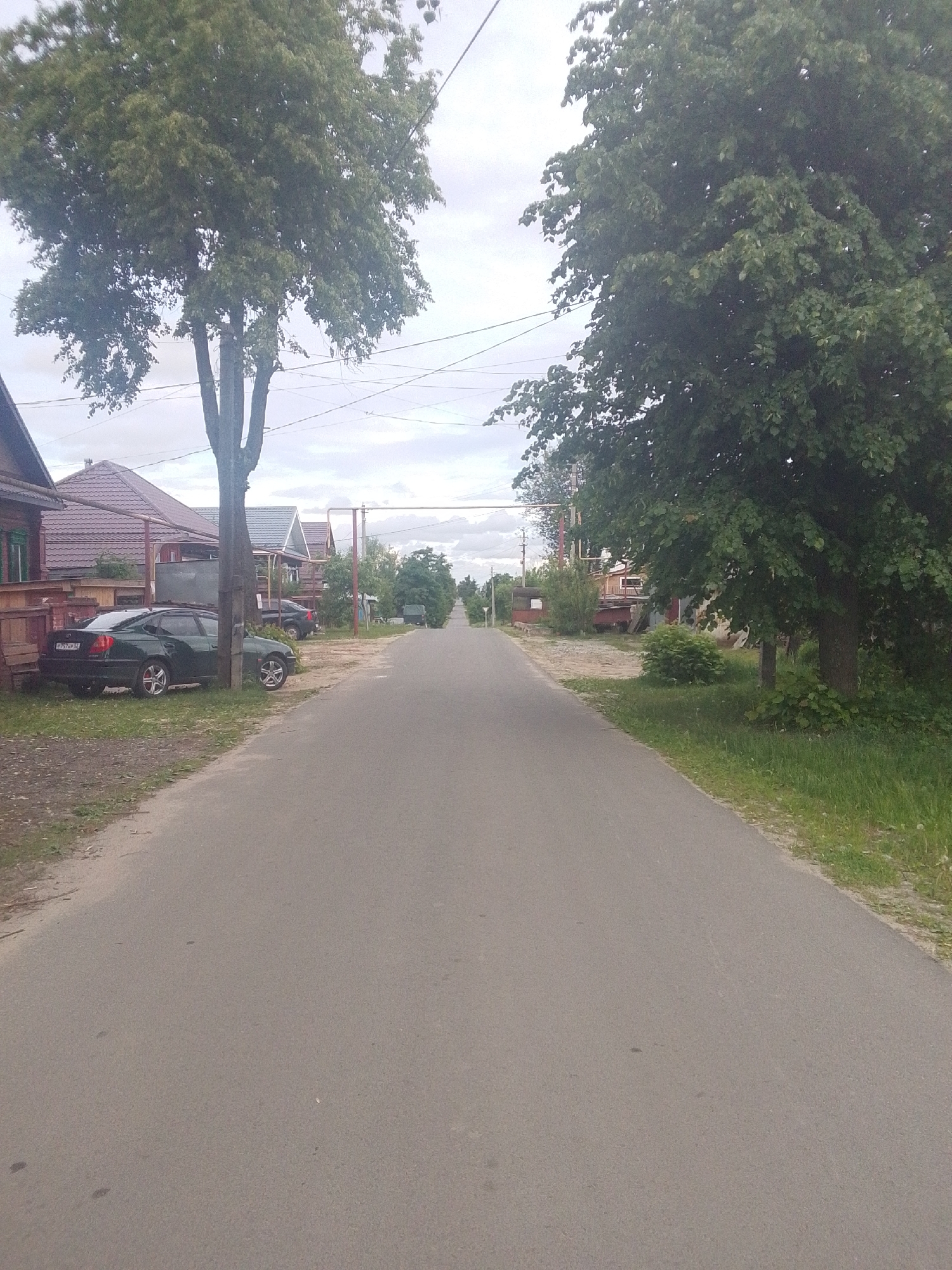 The image shows a street in an area with houses. There is a paved road running through the center of the frame. On either side of the road are trees and grass. Houses are visible behind the trees on the left side of the road, with a car parked in front of one. There are overhead wires running above the street. The location is unknown, Unknown.