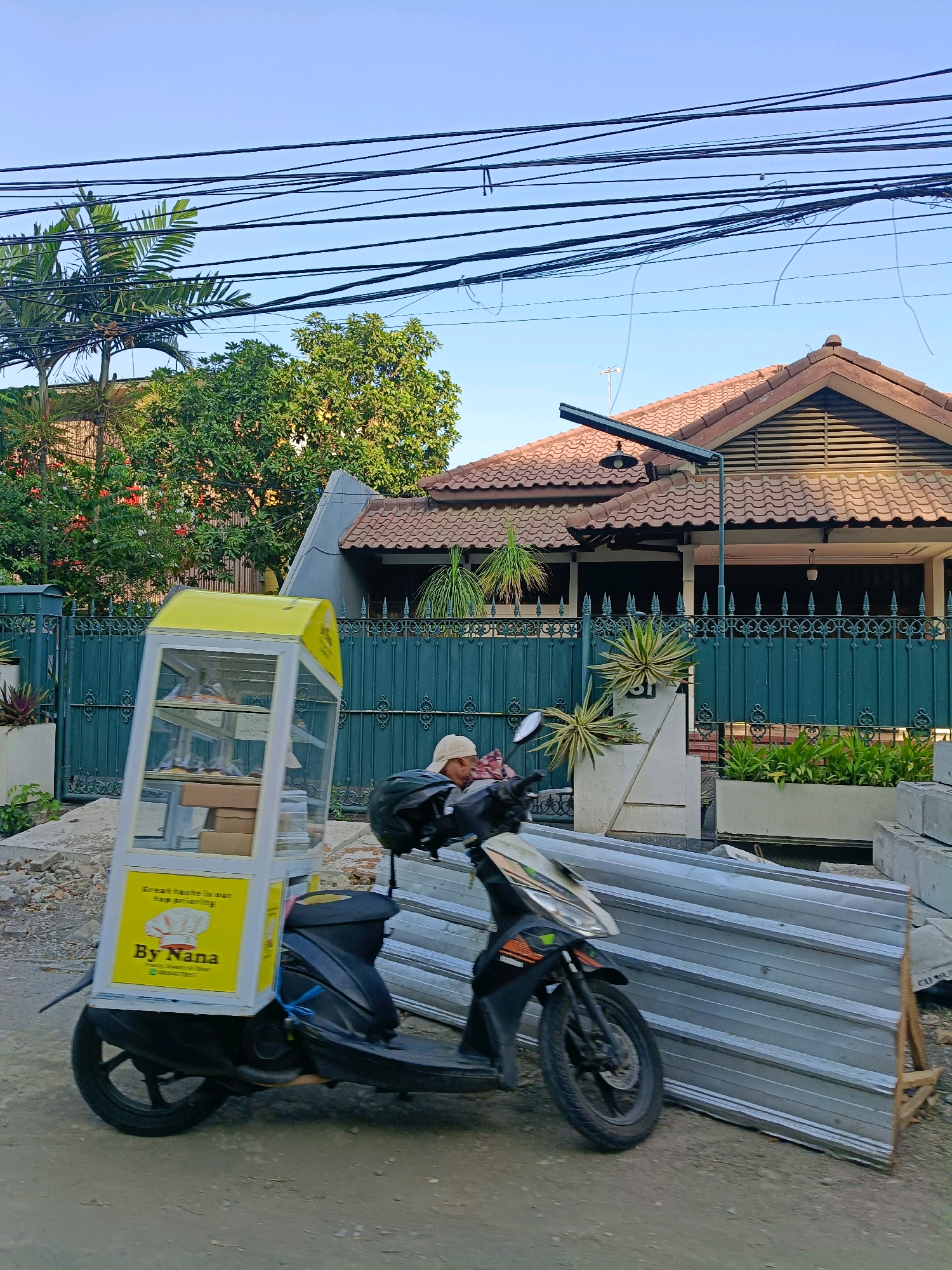A scooter is parked on a dirt road in front of a house with a green metal fence. The scooter has a yellow display case attached to the back, which appears to be a food or product vendor. The display case has "By Nana" written on it. A person wearing a beige cap is seated on the scooter. In the background, there is a house with a tiled roof and a solar-powered light fixture. Several large metal sheets are leaning against a wall to the right of the scooter. Overhead, a tangle of power lines crisscrosses the sky. The scene suggests a residential area in Surabaya, Indonesia, with a street vendor preparing for or concluding business.