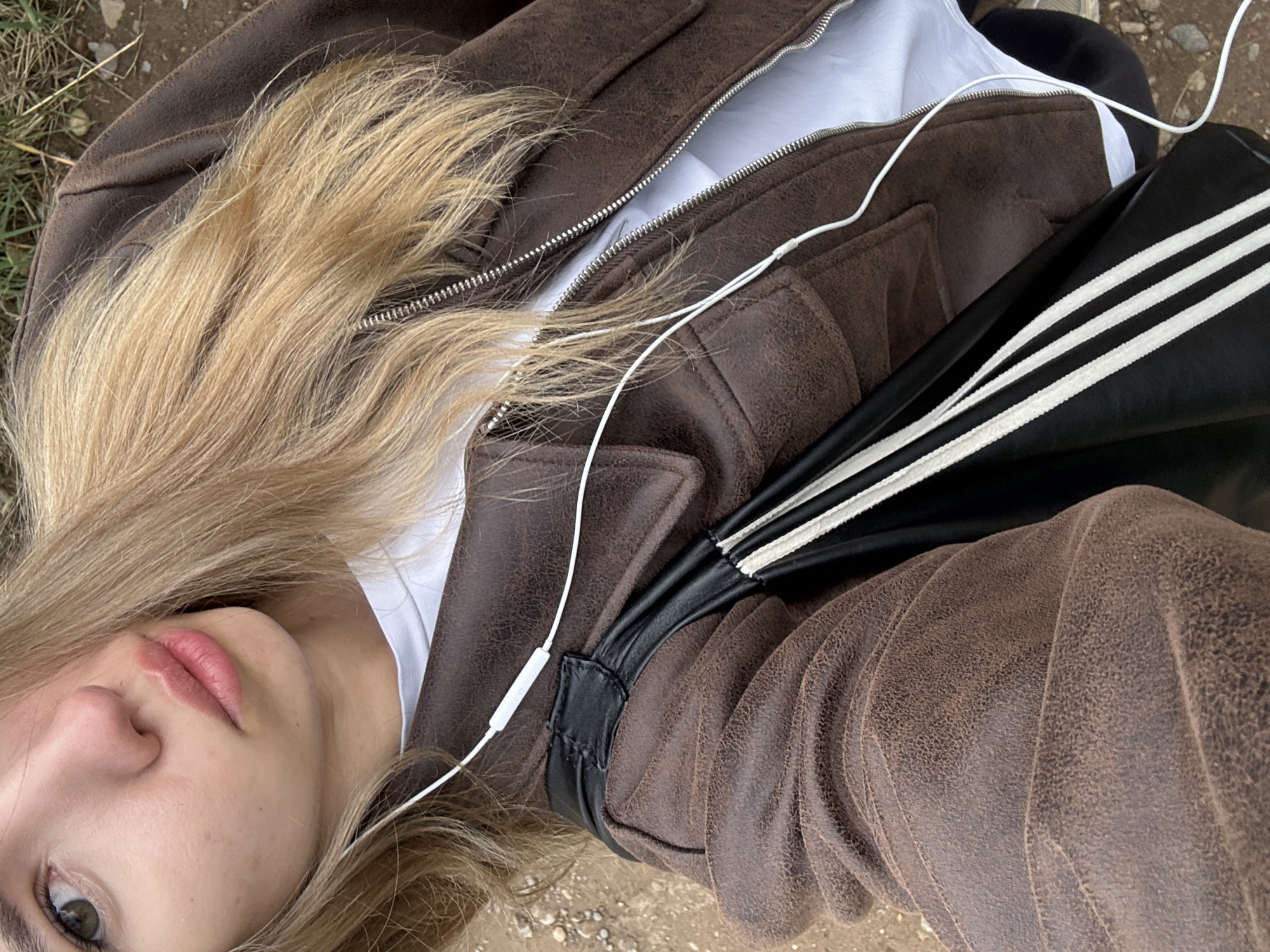 The image shows a close-up, angled view of a person with blond hair, fair skin, and pink lips. They are wearing a brown jacket with a visible white zipper and a white t-shirt underneath. A black bag with three white stripes is partially visible. A pair of white earphones is draped across the jacket. The ground is visible in the background. The image provides no information about the location being Noviy, Russia.