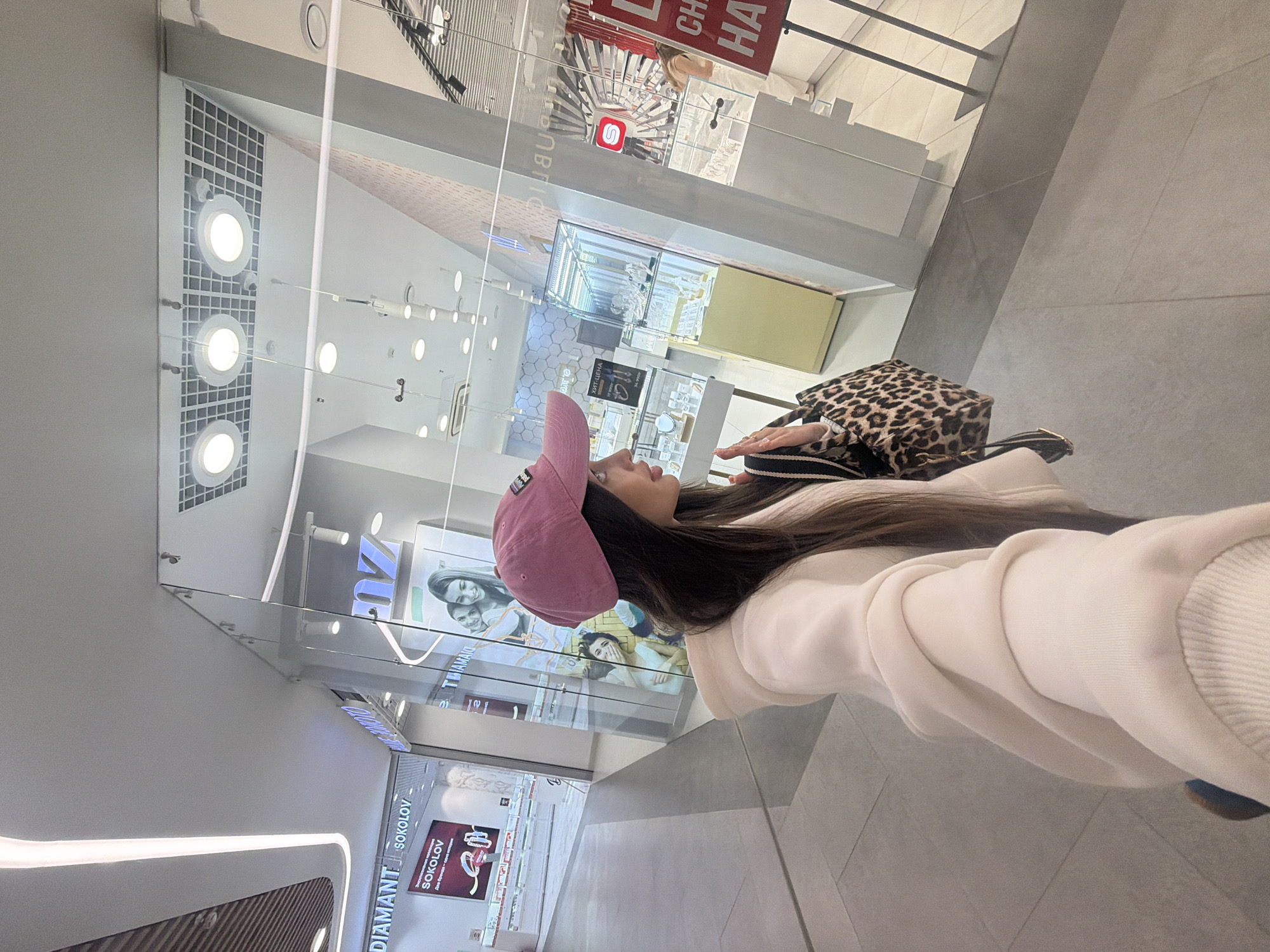 A person is captured in profile within what appears to be a modern shopping mall in Izhevsk, Russia. The individual wears a pink baseball cap, a light-colored hooded sweatshirt, and has long, dark hair. They are holding a distinctive leopard print shoulder bag.
The setting features bright, artificial lighting from numerous circular ceiling lights and linear light strips. The background is dominated by glass storefronts, creating reflections of the interior space. The floor is covered with large, light-grey tiles.
Visible signs and text include "DIAMANT" and "SOKOLOV" in both Latin and Cyrillic script, identifying a jewelry store. Other Cyrillic text is seen on various signs, including "ЮВЕЛИРНЫЙ" (Jewelry) and large red letters spelling out "СКИДКИ" (DISCOUNTS) and partially "ДО ХАЛЫ". One storefront displays a large image of two smiling women. Another features a stylized red 'S' logo. The reflections in the glass show more store interiors with jewelry displays.
The person appears to be standing or casually walking, observing the surroundings of the mall. The time of day is not evident due to the indoor lighting. The prominent display of Russian brands and Cyrillic text confirms the location context of Izhevsk, Russia.