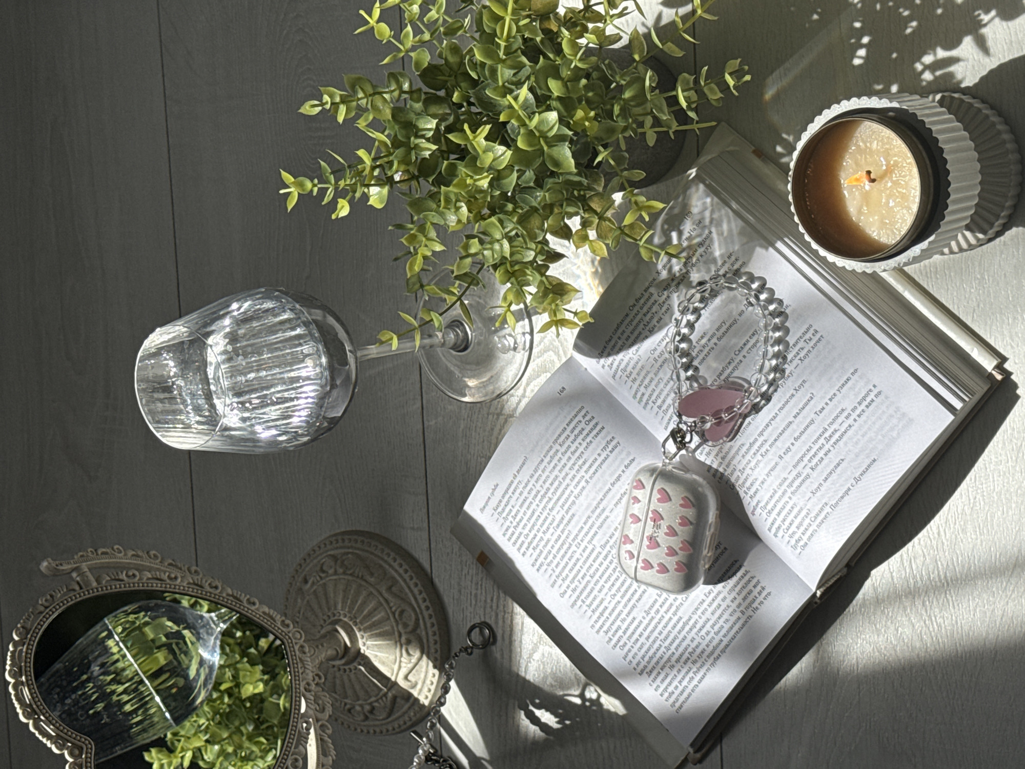 A flat lay scene on a light-colored wooden surface captures a still life arrangement bathed in soft sunlight. In the upper left, a vintage heart-shaped mirror with ornate detailing reflects a glass of water and a small green plant. To its right, a clear stemmed wine glass, filled with water, sits prominently. In the foreground, an open book lies flat, its pages partially obscured by an open AirPods case adorned with small pink hearts. Attached to the AirPods case is a silver beaded bracelet with a pink heart charm. To the right of the book, a small potted plant with lush green leaves is positioned near the wine glass. In the lower right corner, a lit candle in a ribbed white holder emanates a warm glow. The overall impression is one of peaceful contemplation and aesthetic arrangement, with no people or identifiable urban elements present.