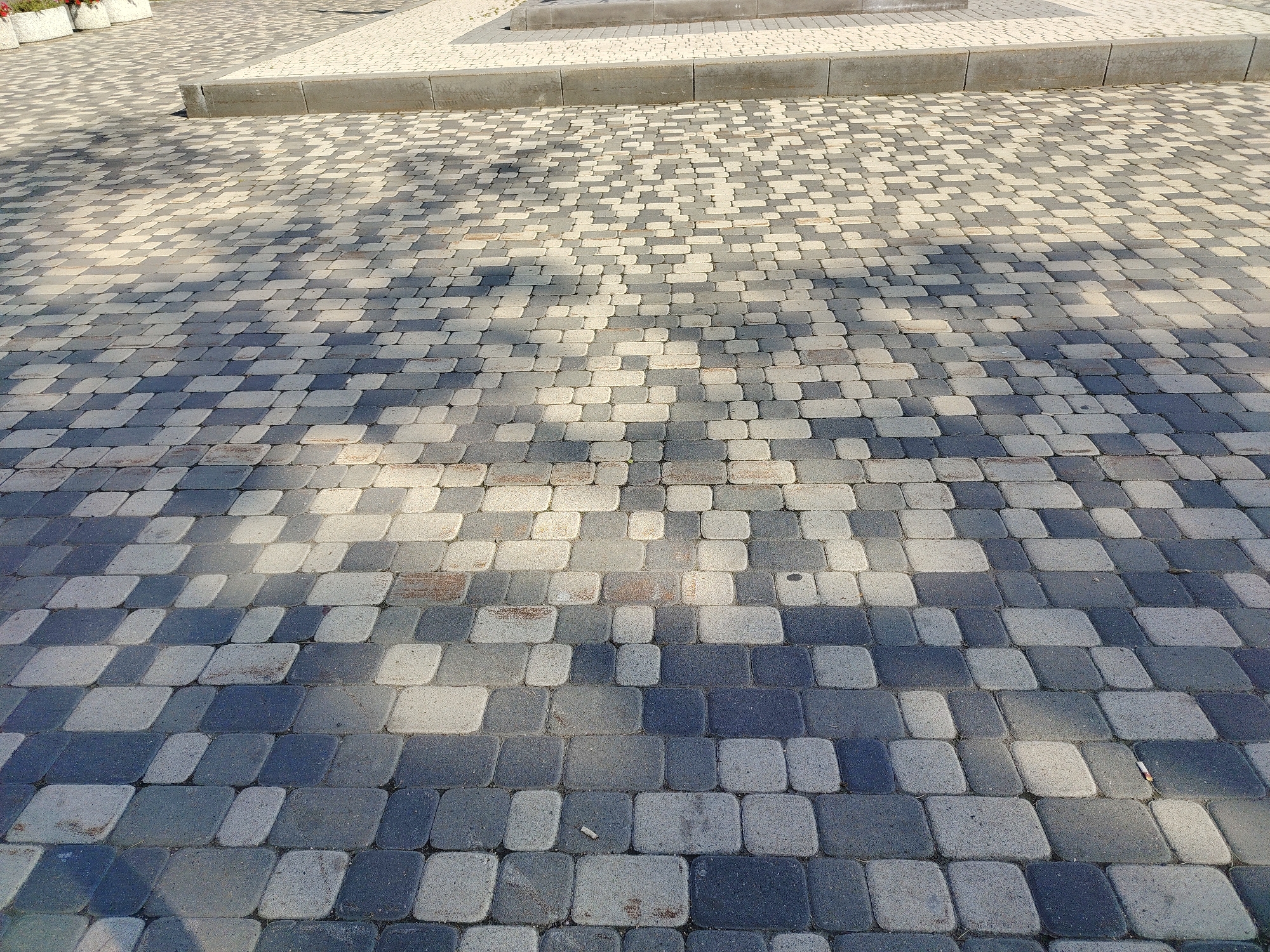 The image depicts a pedestrian walkway paved with grey and black interlocking concrete pavers. The pavers are laid in a pattern of alternating colors and orientations, creating a textured, visually dynamic surface. Sunlight casts distinct shadows across the pavement, highlighting the three-dimensional nature of the paving stones and creating a dappled effect. The walkway appears to be part of a public space, possibly a park or a plaza, given the presence of a concrete curb and what appears to be a planter box in the upper left corner of the image. The scene is devoid of people. The location is identified as Kemerovo, Russia.