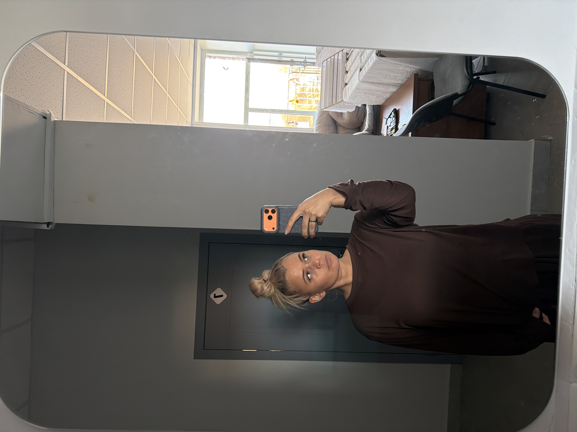 The image displays a mirror selfie taken by a woman indoors, with the photograph itself oriented sideways. The woman, with blonde hair styled in a bun, is shown from the chest up, wearing a long-sleeved, brown, loose-fitting top or dress. She holds a dark smartphone, featuring an orange accent around its camera module, to capture her reflection. Her expression is neutral, and she appears to be looking slightly past the camera. Notable details include multiple ear piercings and a ring on her left hand.
Behind the woman's reflection, a hallway or corridor is visible. This includes a plain grey wall leading to a dark grey door and its frame. On the door, a white square symbol with a black border contains the number "1" and an arrow pointing to the right.
Above the wall and door in the mirror's reflection, a large window reveals a brightly lit outdoor scene, indicating daytime. Parts of building exteriors are visible through the window. To the right of this window reflection, an indoor space is partially seen, featuring a table covered with a white cloth and a black chair. The upper left portion of the mirror reflects a white acoustic tile ceiling with a grid pattern.
The setting appears to be an indoor environment, likely a commercial or institutional building, or an apartment hallway, potentially in Sarapul, Russia. The scene is well-lit by natural light from the reflected window.